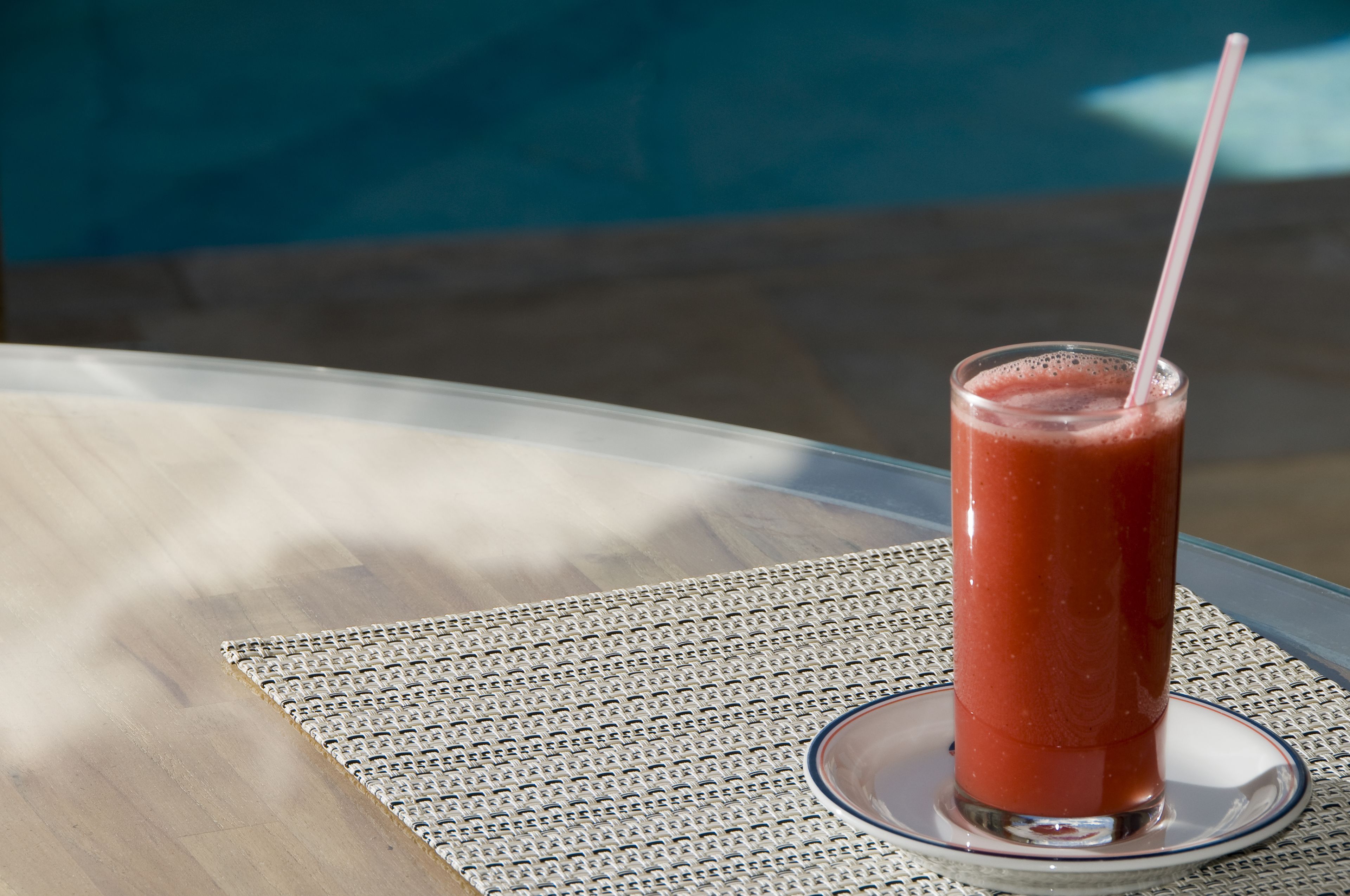 A glass of red beverage on a table with a woven placemat, near a pool.