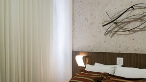Habitación de hotel con cama, cortinas blancas, aire acondicionado de pared y mesita de noche con lámpara.