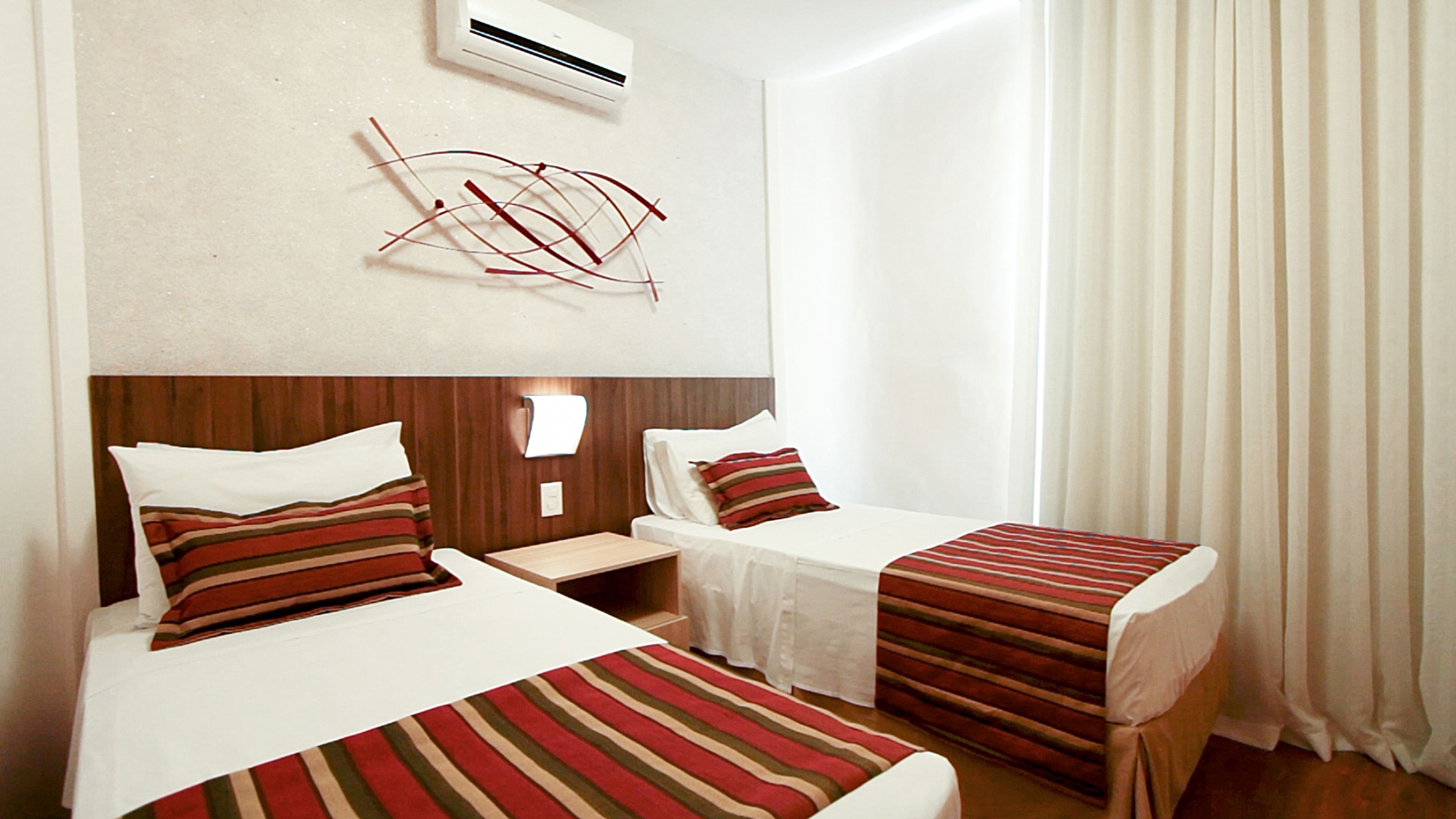 A hotel room with two beds, each with striped bedding, a wooden headboard, a bedside table, and a wall-mounted air conditioner.
