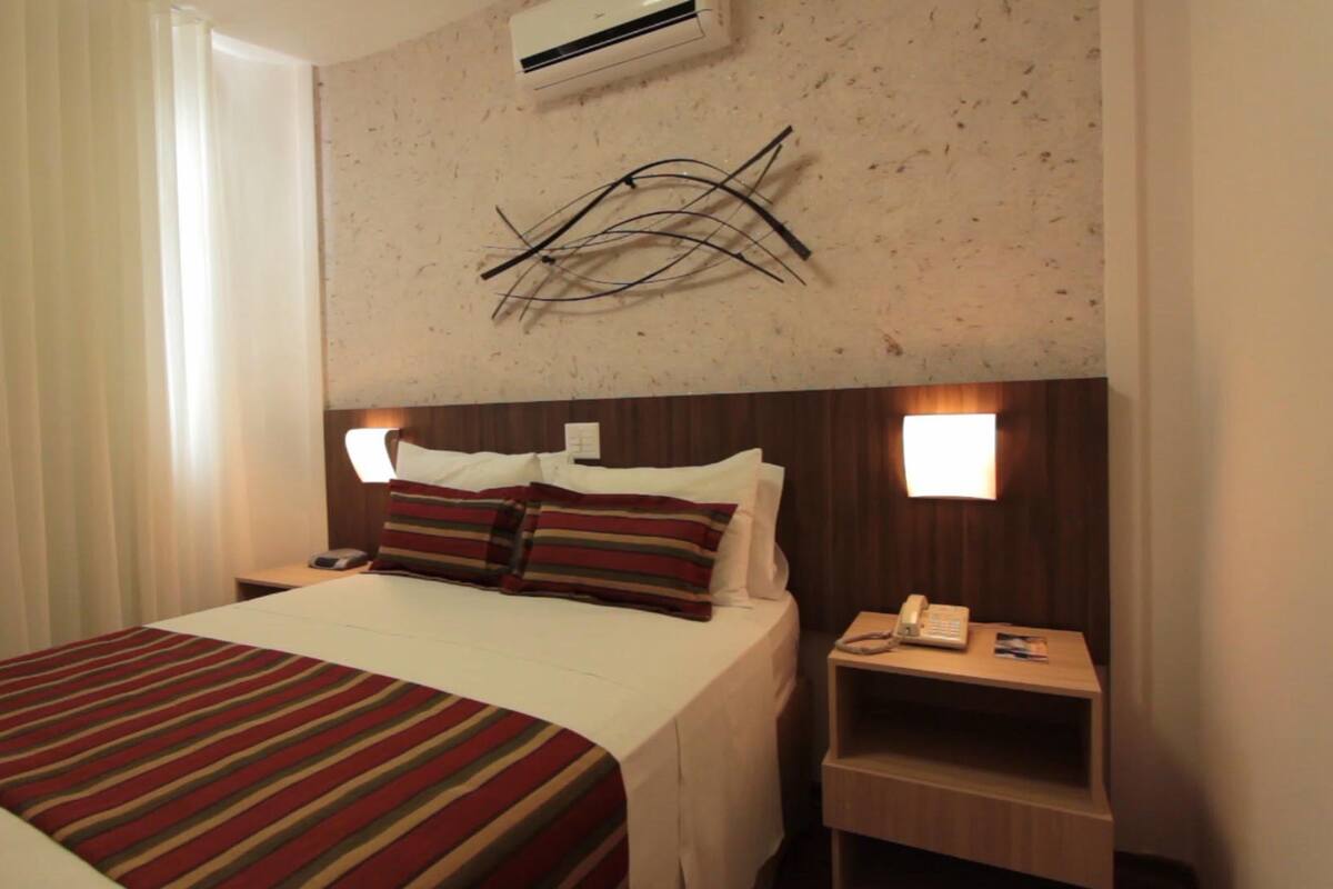 Een hotelkamer met een bed, een nachtkastje met telefoon en een wandairconditioner.
