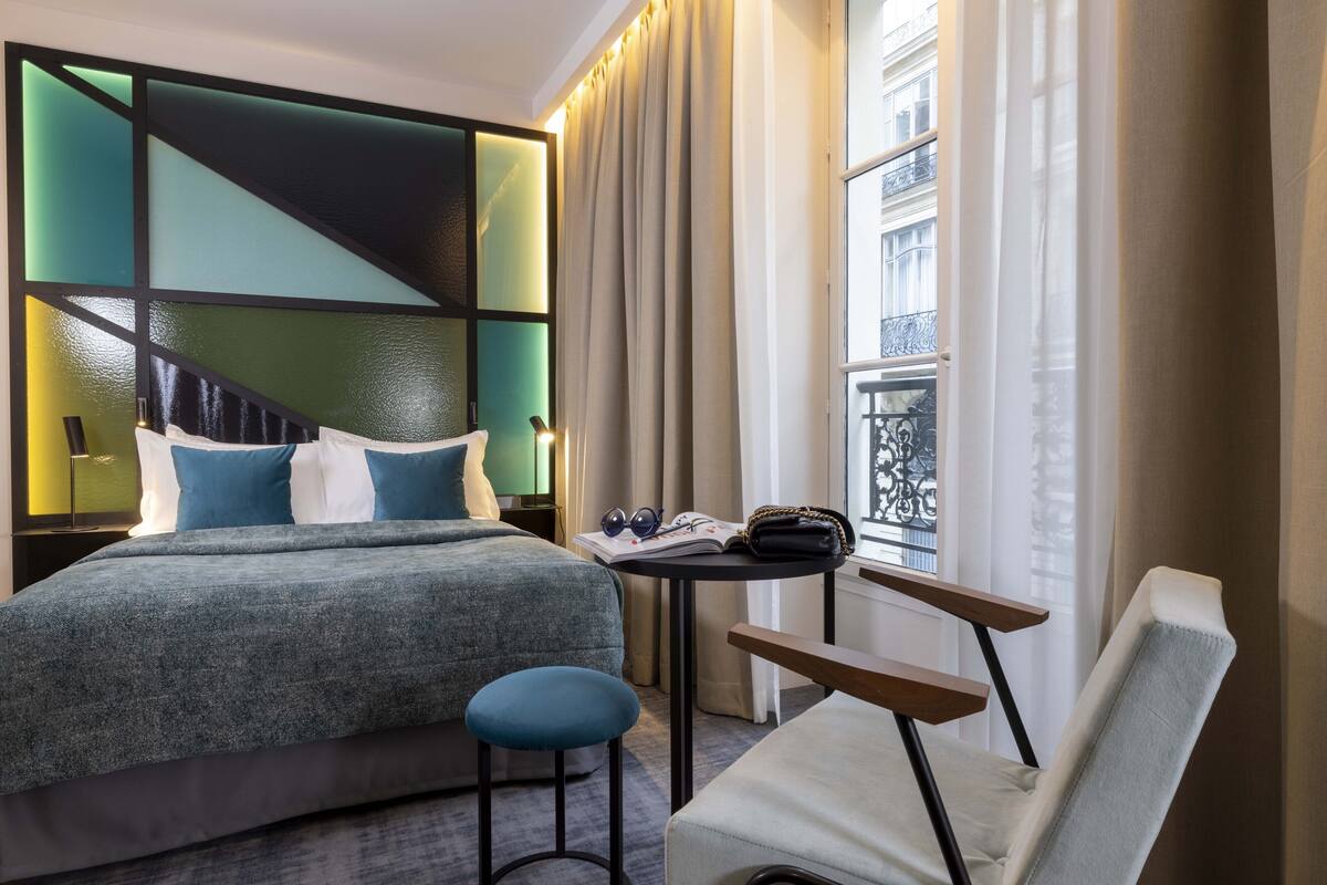 A hotel room with a bed, a small table with a chair, and a view of the city through large windows.