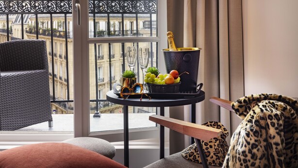 A balcony with a wicker chair, a table with a champagne bottle, glasses, and fruit. - Le 12 Hotel (Paris)