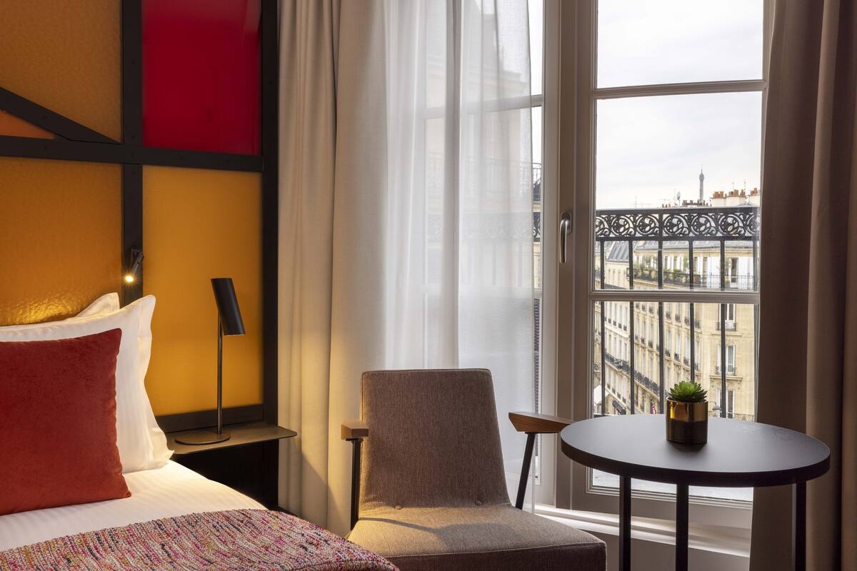 A hotel room with a bed, a chair, a small table, and a view of the city through large windows.