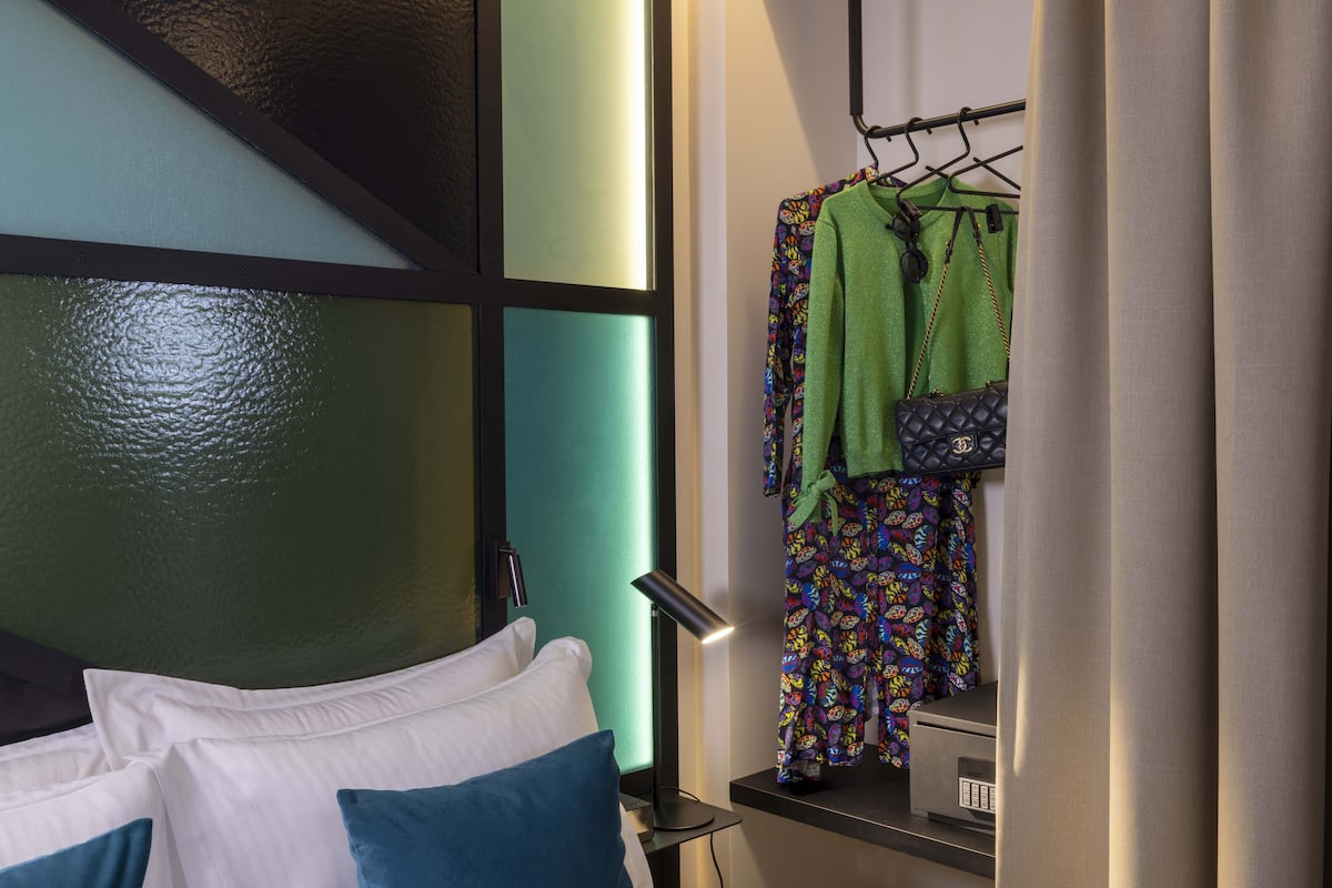 A hotel room with a bed, a nightstand, a lamp, and a clothing rack with a green garment and a patterned outfit.