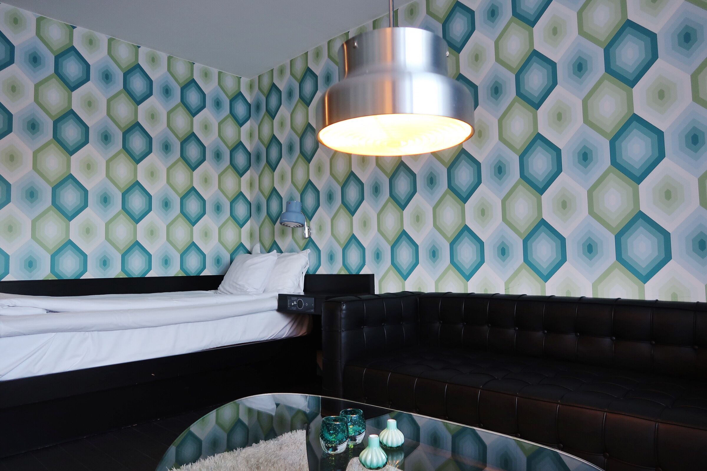 A hotel room with a bed, a black leather sofa, a glass coffee table with decorative items, and a patterned wallpaper.
