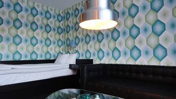 A hotel room with a bed, a black leather sofa, a glass coffee table with decorative items, and a patterned wallpaper.