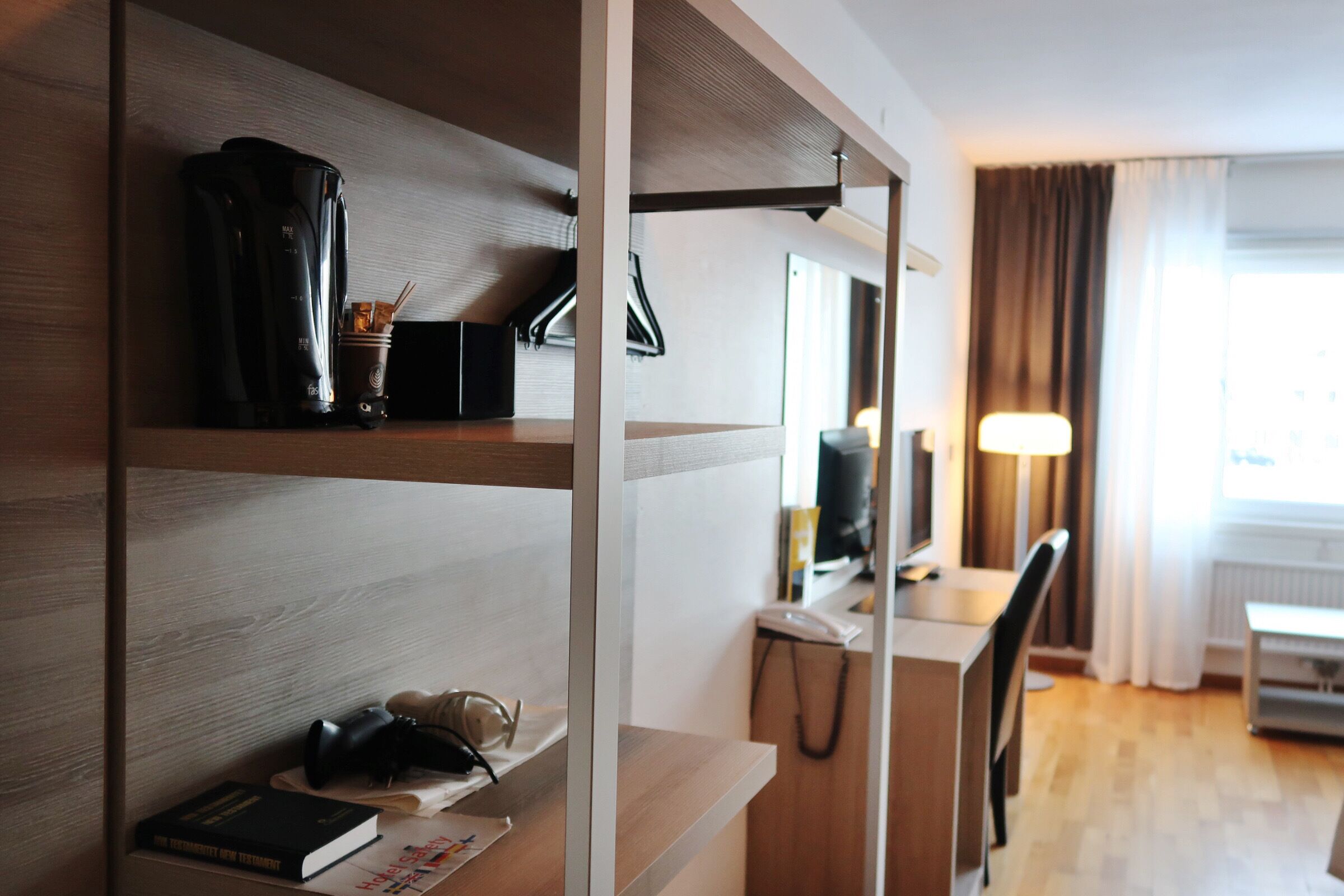 Habitación de hotel con un escritorio de madera, una silla, una lámpara, una cafetera y una estantería con libros y un par de gafas.