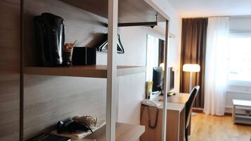 Habitación de hotel con un escritorio de madera, una silla, una lámpara, una cafetera y una estantería con libros y un par de gafas.