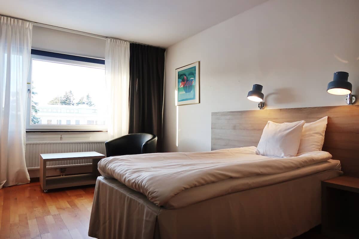 A hotel room with a bed, bedside table, chair, and a window with curtains.