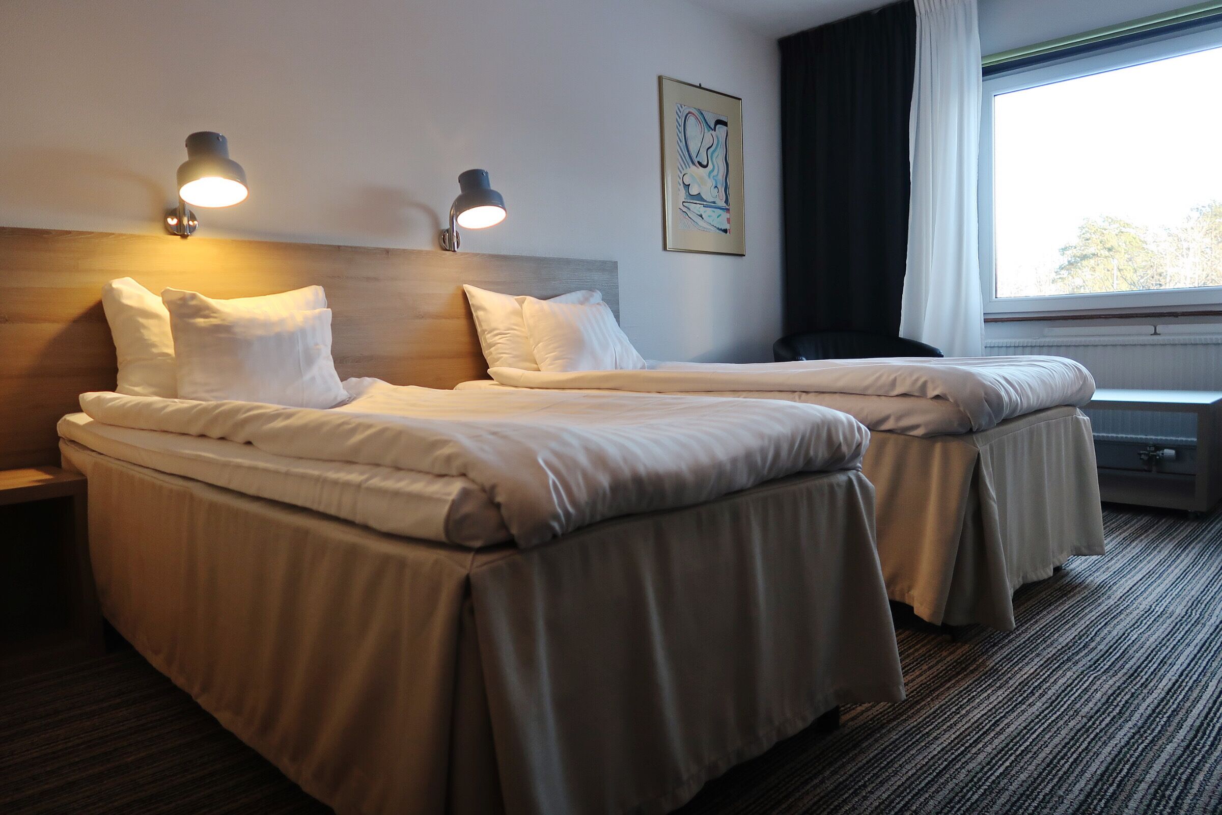Een hotelkamer met twee bedden, een hoofdbord, wandlampen, een raam met gordijnen en een kunstwerk in een lijst aan de muur.