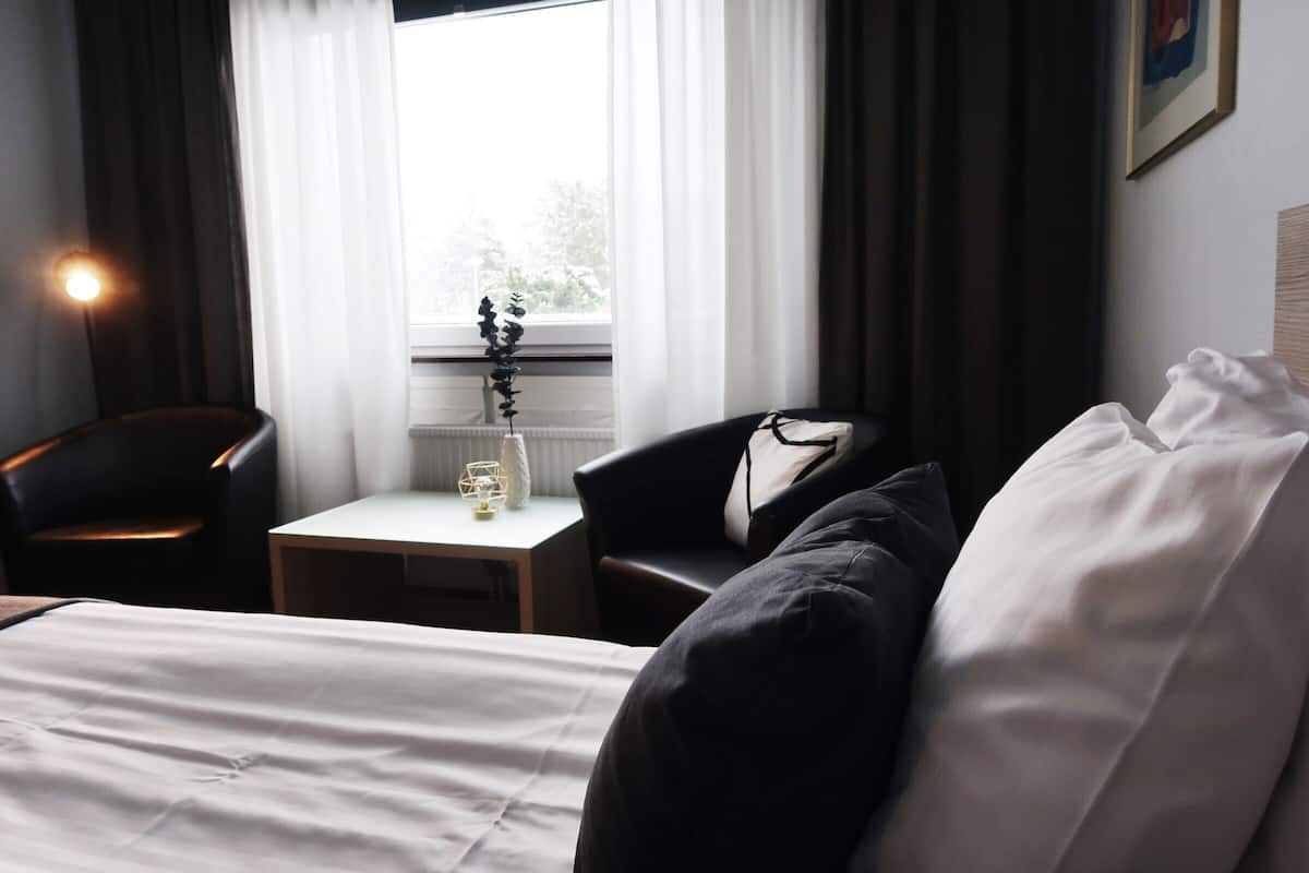 A hotel room with a bed, a chair, a small table with a vase, and a window with curtains.