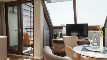 Una habitación de hotel moderna con un ventanal grande, un televisor de pantalla plana, una mesa de comedor con sillas, un sofá y un balcón con una silla.