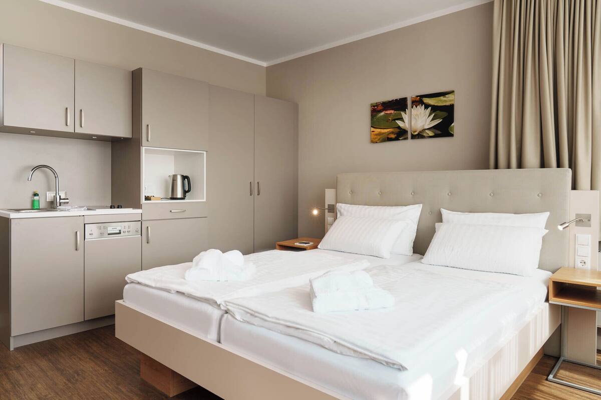 A hotel room with a bed, a nightstand, a bedside table, a wardrobe, a kitchenette, and a bathroom.
