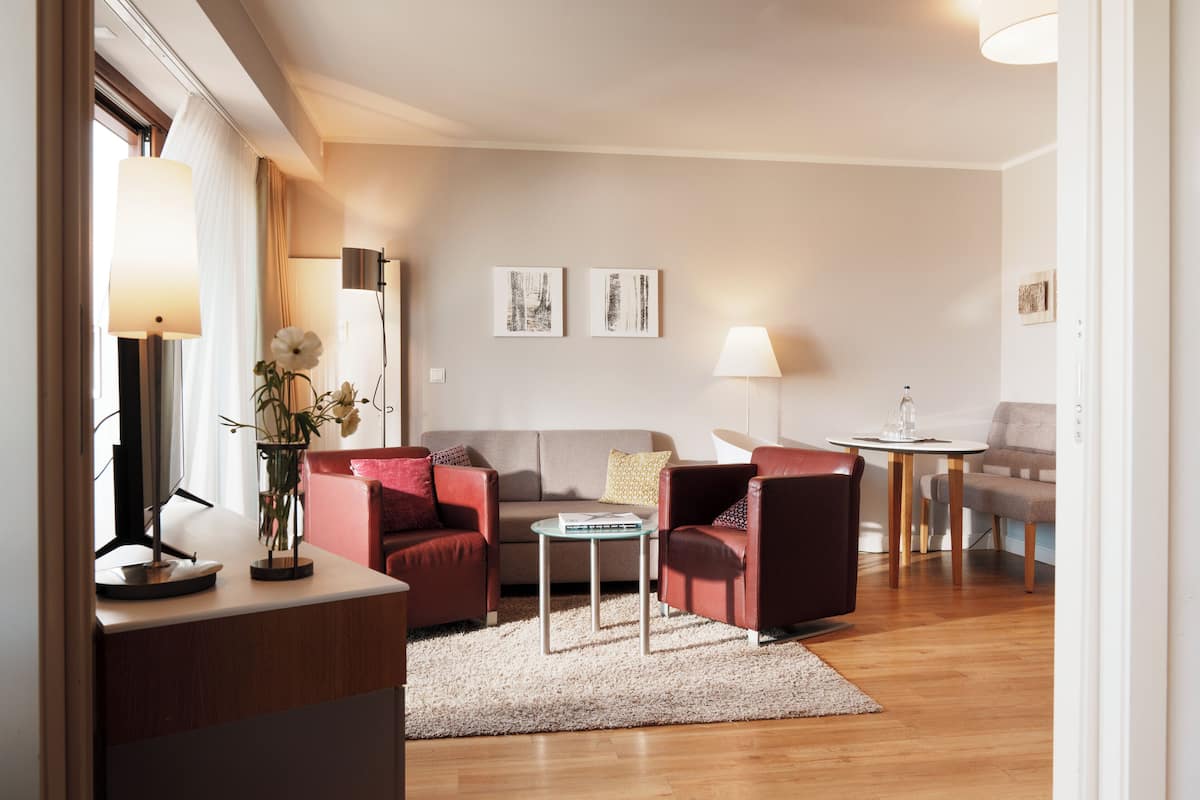 A modern living room with a sofa, armchairs, a coffee table, and a lamp. There are two framed artworks on the wall and a vase with flowers on the coffee table.