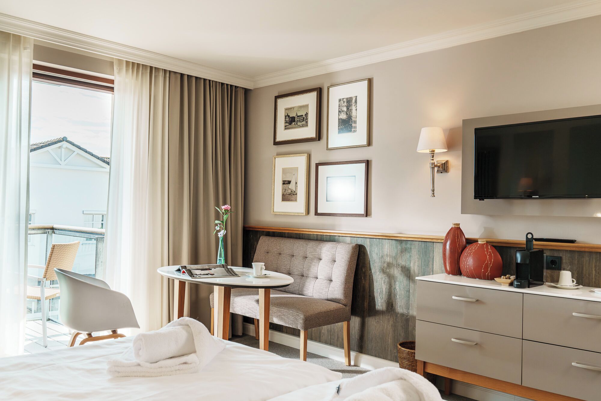 A hotel room with a bed, a chair, a small table with a cup, a TV, and framed pictures on the wall.