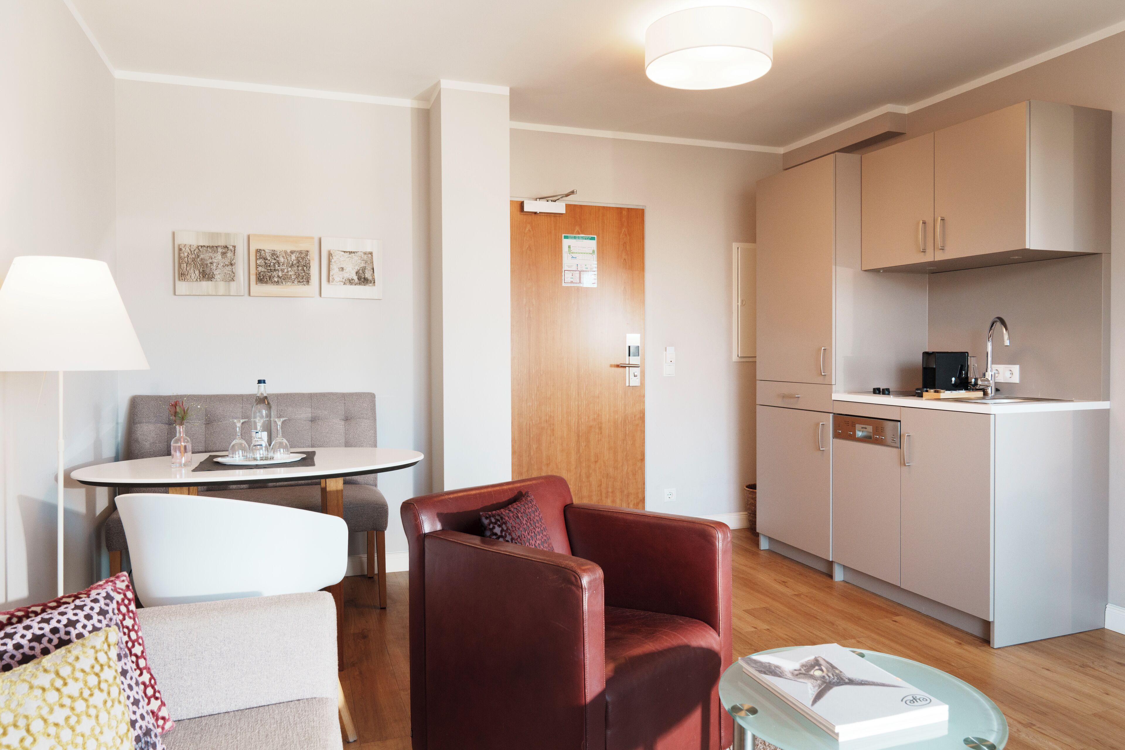 A modern living room with a sofa, armchair, and dining table. There is a kitchen area with cabinets and a countertop.