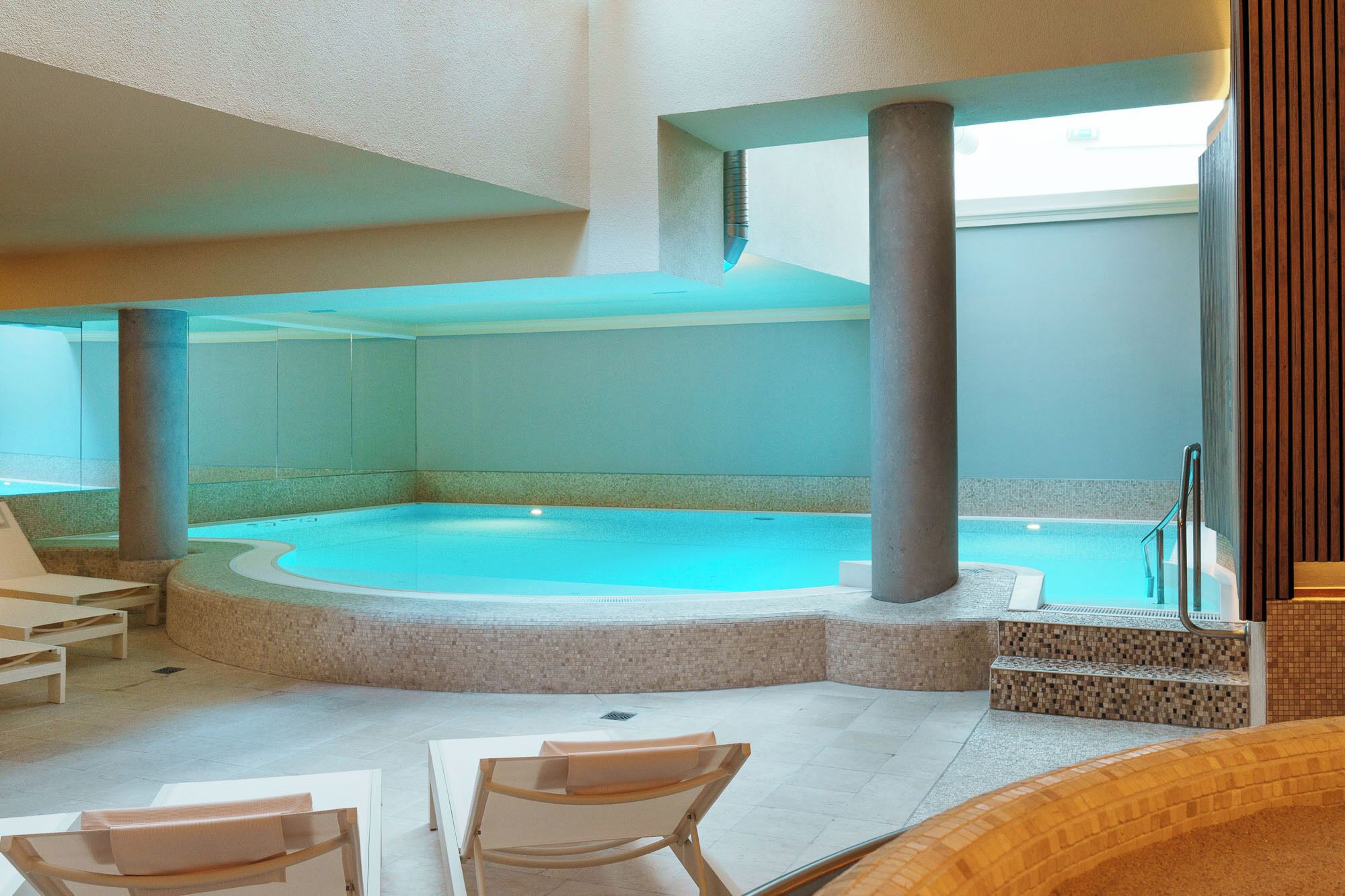 Una amplia piscina interior con borde de piedra, rodeada de zonas de estar y suelo de baldosas.