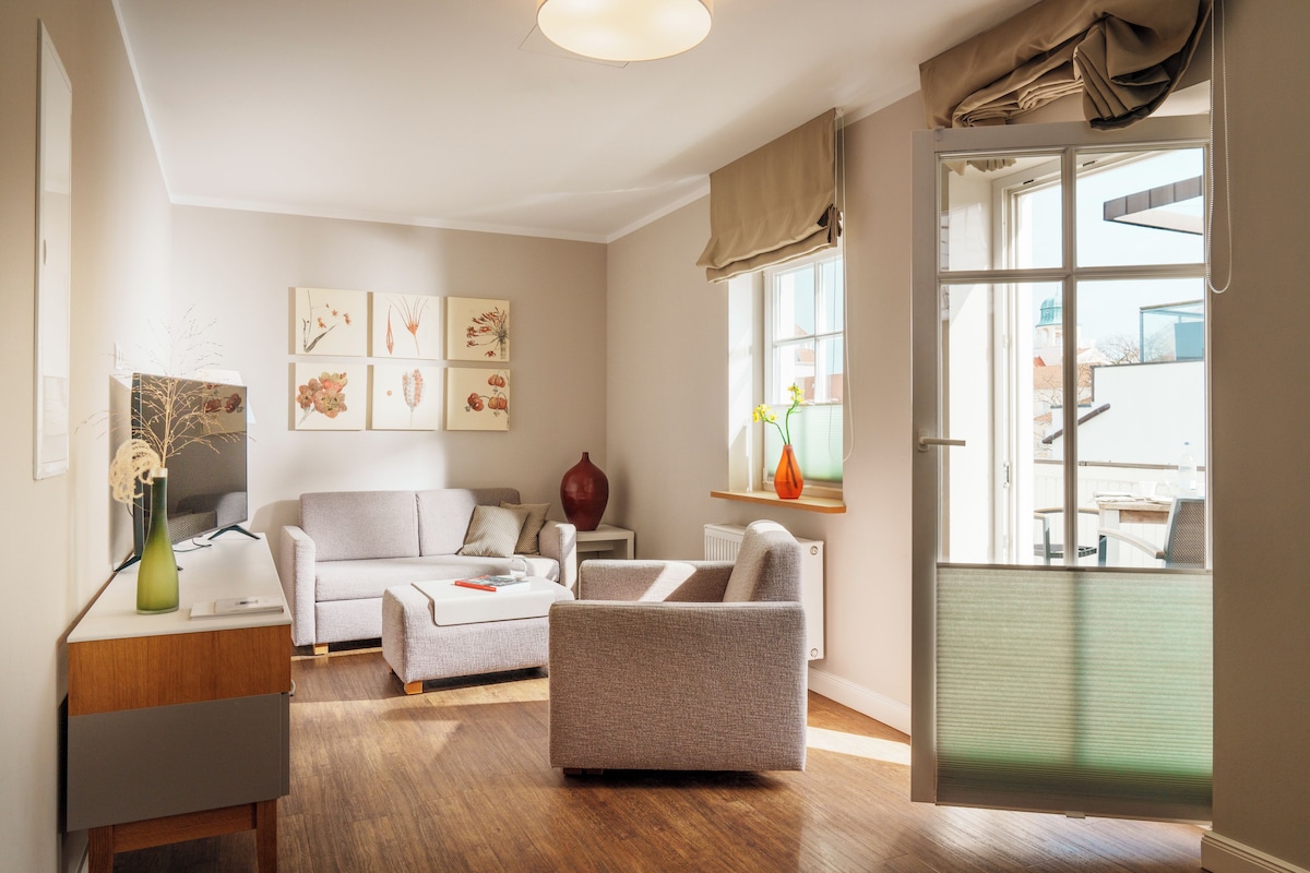 A modern living room with a sofa, armchair, and a coffee table. There is a vase on a side table and artwork on the wall.