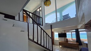 Ein geräumiges Zimmer mit einer Treppe zu einer oberen Ebene, einem großen Fenster mit Meerblick und einem an der Wand befestigten Flachbildfernseher.