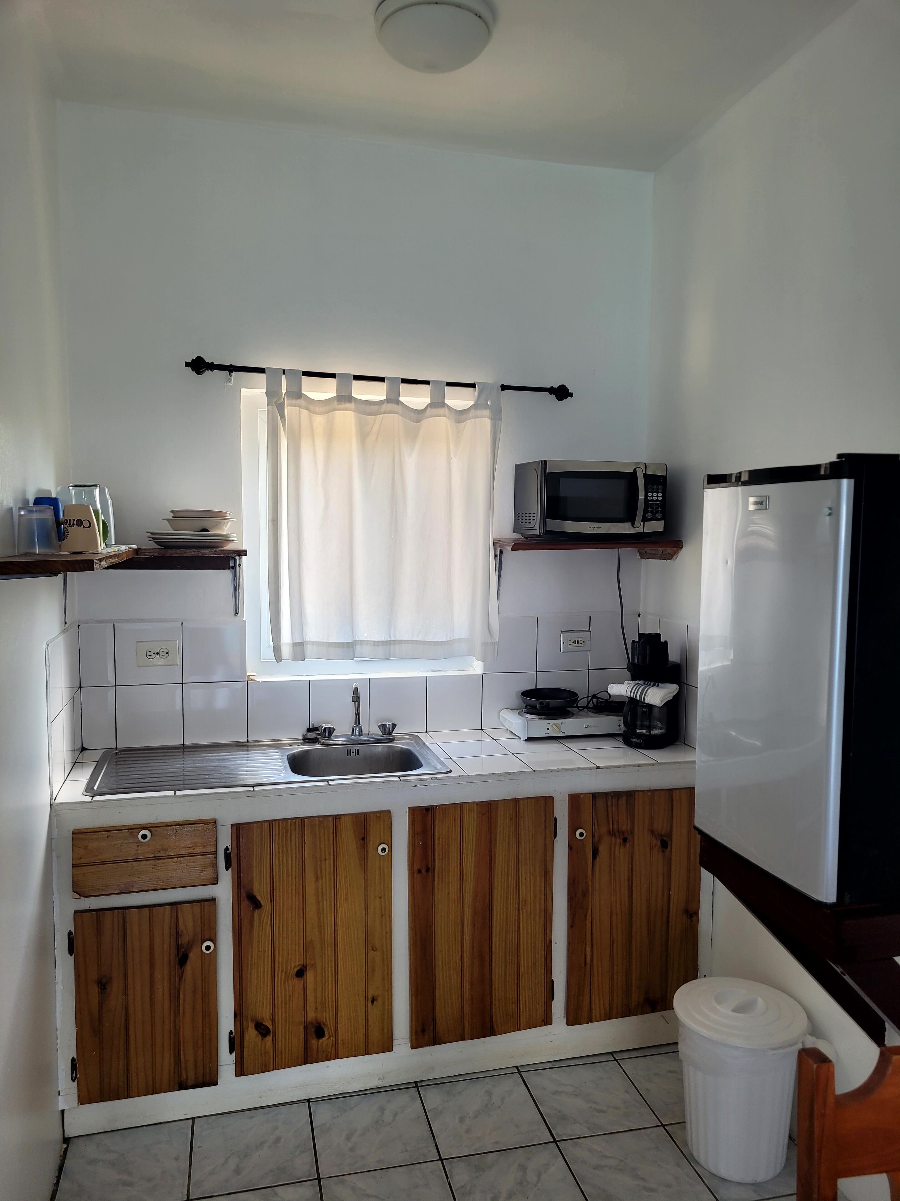 Uma cozinha compacta com azulejos brancos, armários de madeira, uma pia de aço inoxidável, um micro-ondas, uma geladeira e uma janela com cortina branca.