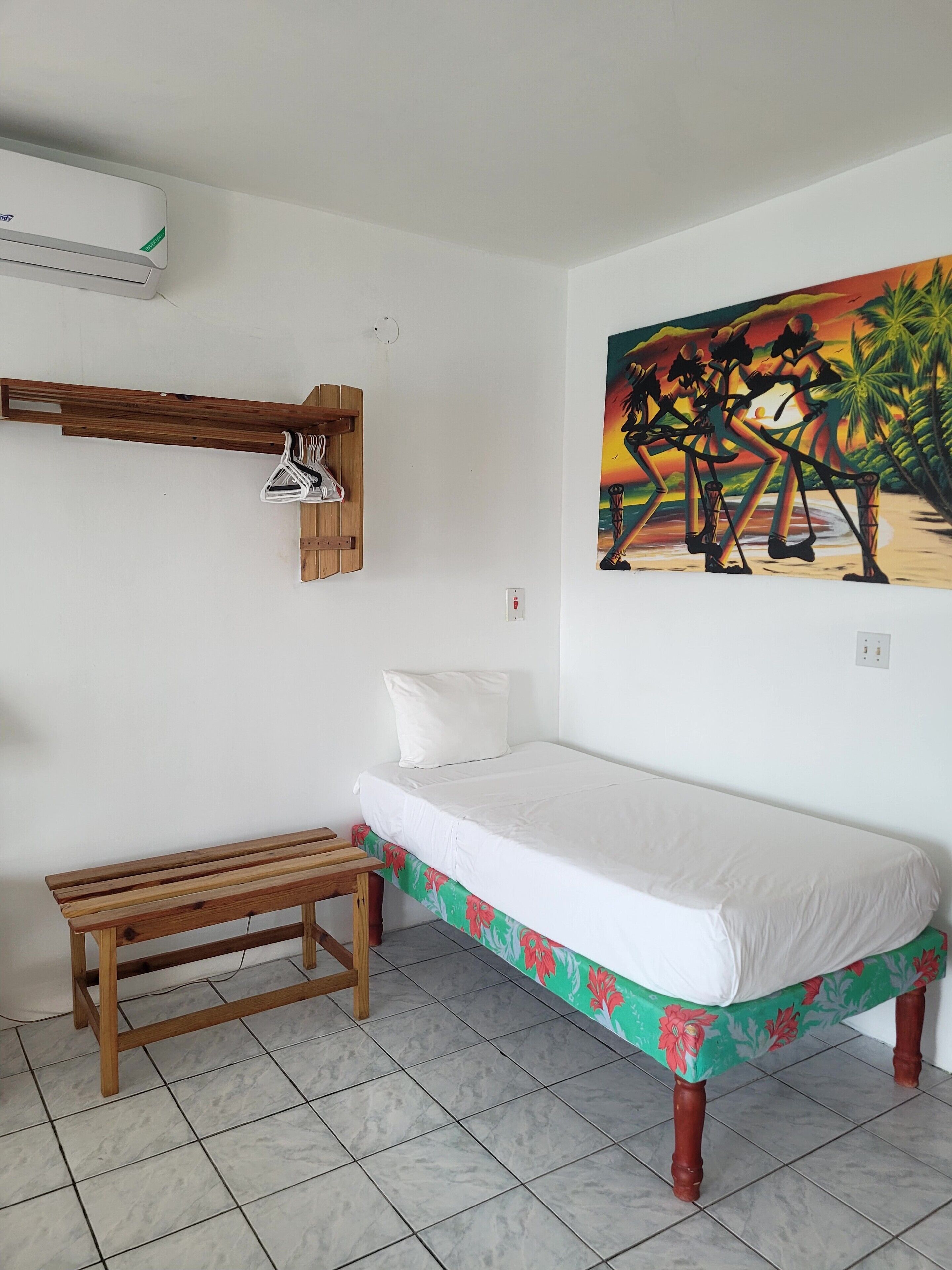 A room with a bed, a wooden bench, a wall-mounted air conditioner, and a painting.