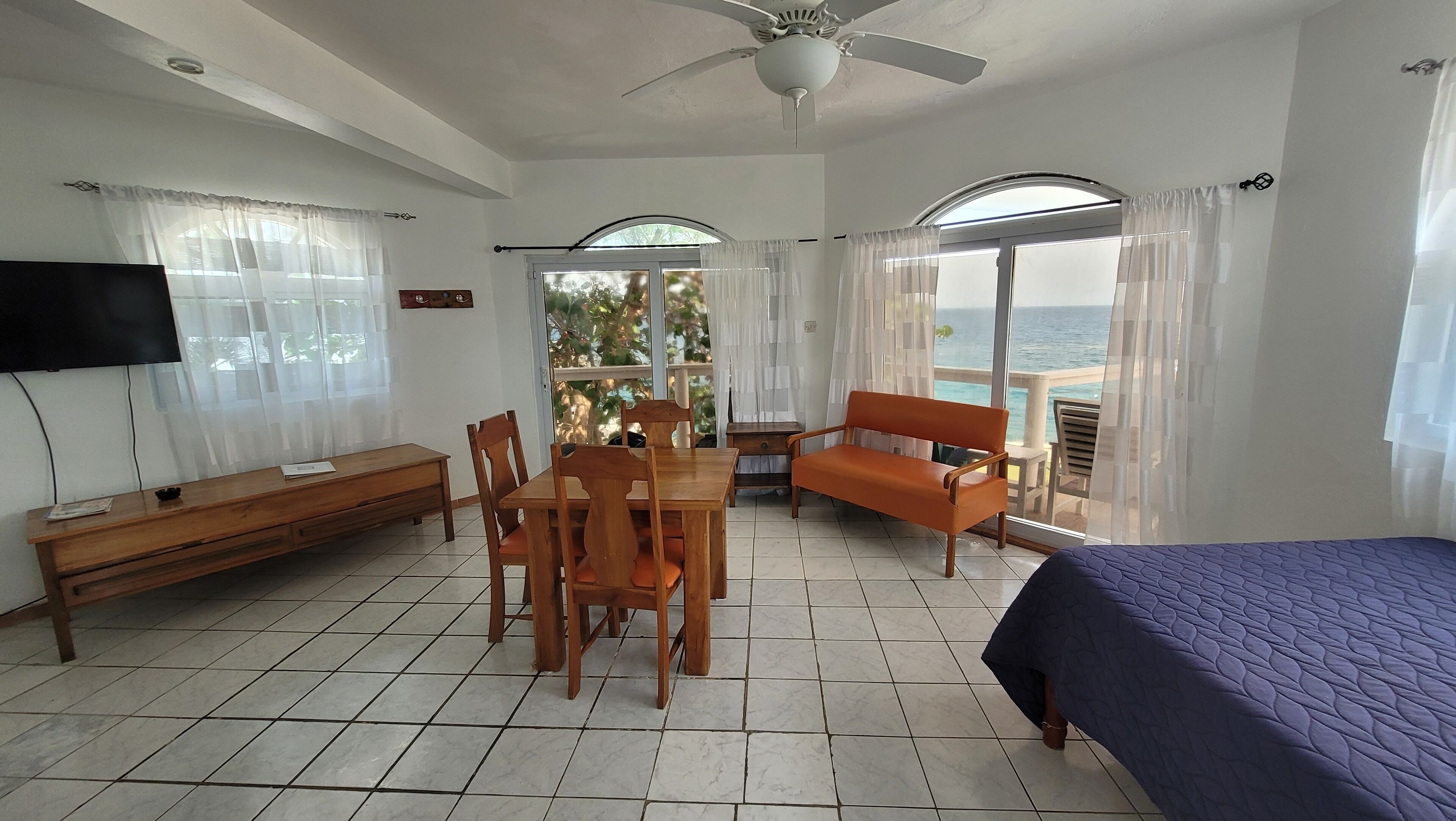 A room with a bed, a TV, a dining table with chairs, a sofa, and a view of the sea.