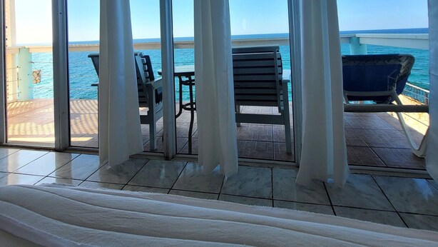 Ein Zimmer mit großen Glasfenstern, die auf eine Terrasse mit Meerblick führen. Auf der Terrasse befinden sich zwei Stühle und ein Tisch.