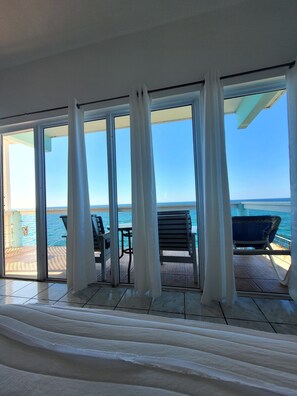 A room with large glass doors leading to a deck with a view of the sea. There are two chairs and a table on the deck. - Home Sweet Home Resort (Negril)