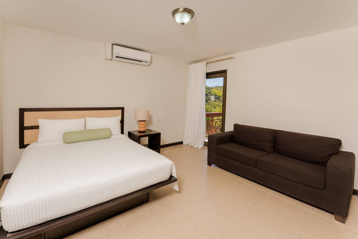 A hotel room with a bed, a brown sofa, a nightstand with a lamp, and a window with a view of greenery.