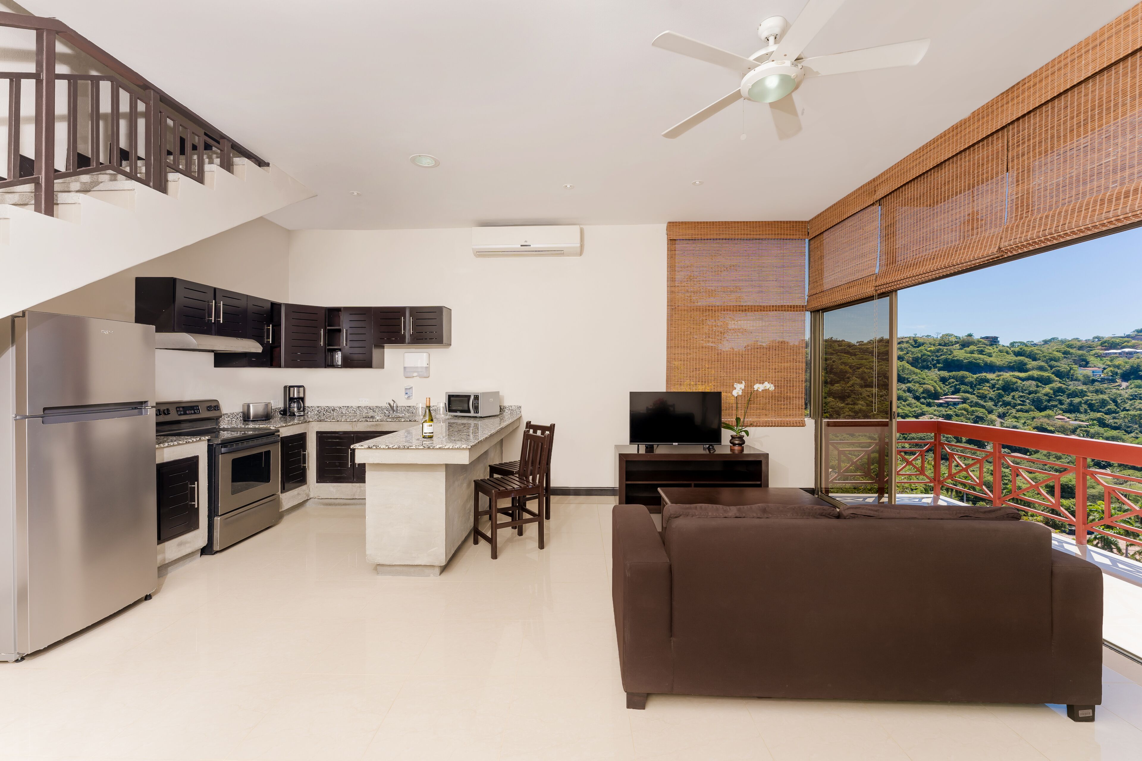 Cozinha moderna com área de jantar, sofá e varanda com guarda-corrimão vermelho com vista para uma paisagem verde.