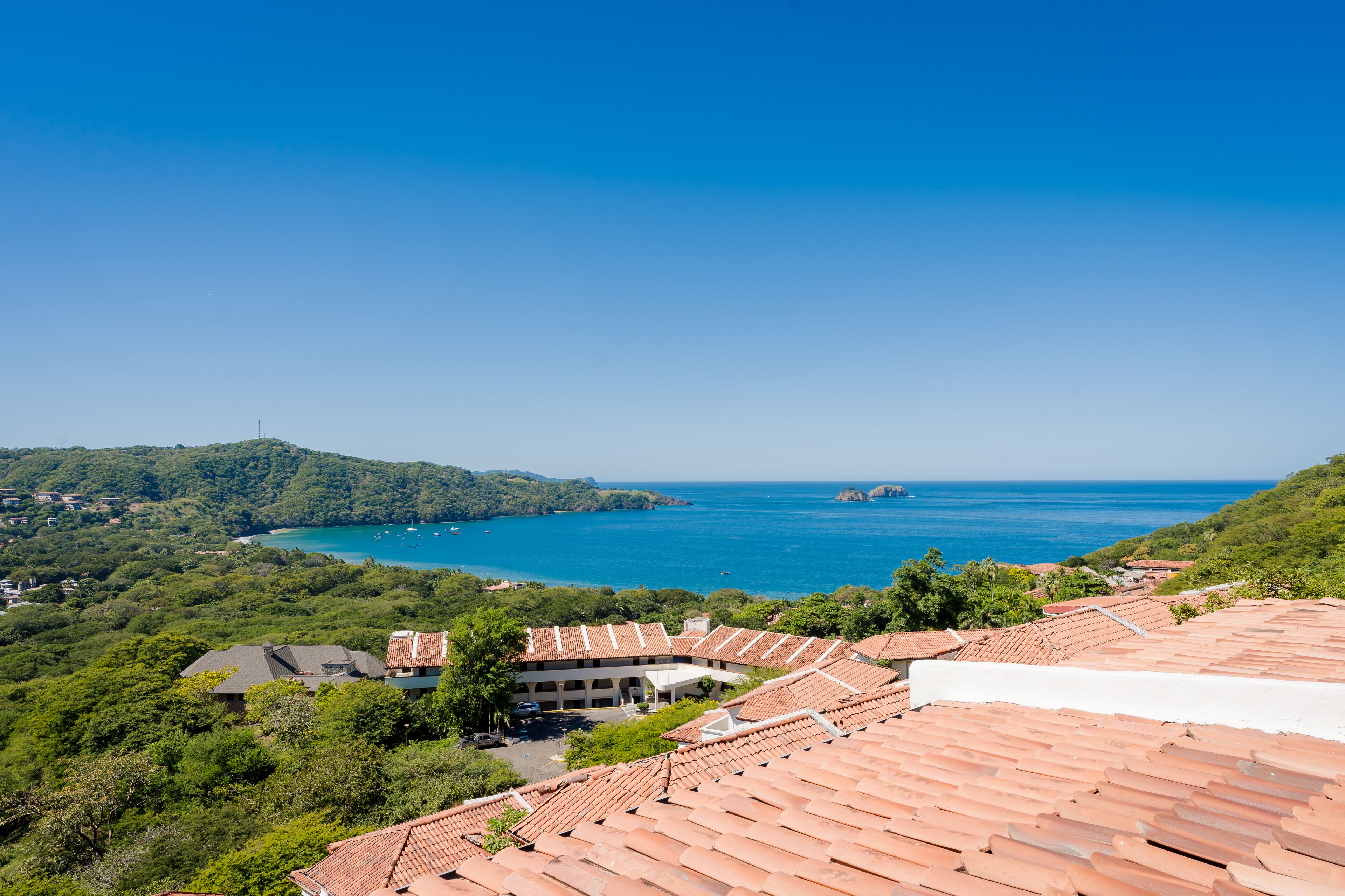 Vista costera con edificios de tejados terracota, abundante vegetación y un mar azul cristalino.