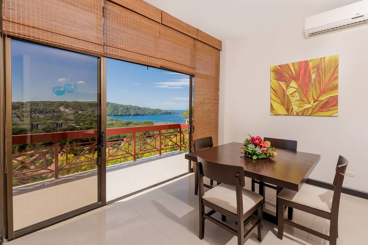 A balcony with a view of a lake and hills, a dining table with chairs, and a wall-mounted air conditioner.