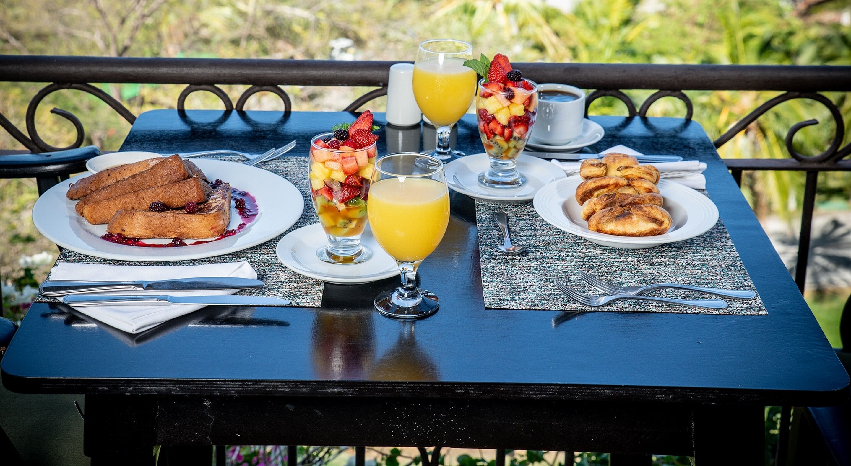 A table set with breakfast items including toast, fruit salad, orange juice, and pastries, outdoors with greenery in the background.
