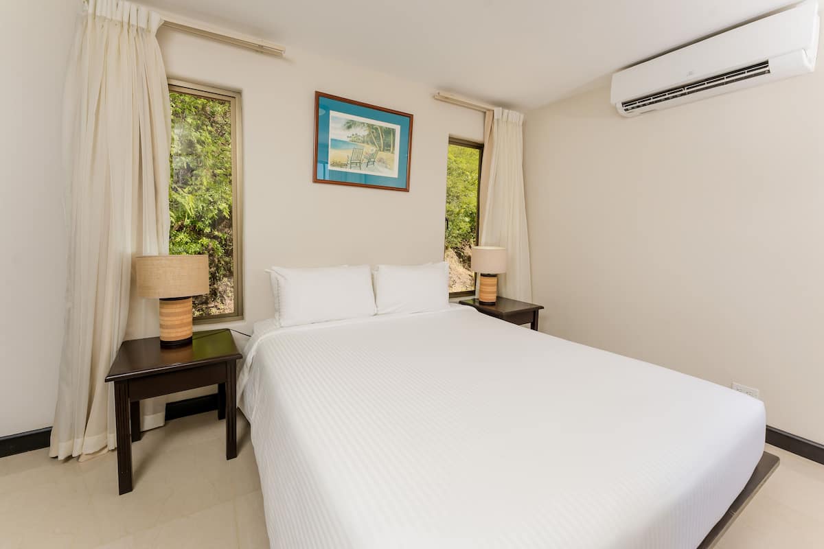 A hotel room with a large bed, two bedside tables with lamps, a window with curtains, an air conditioning unit, and a framed picture on the wall.