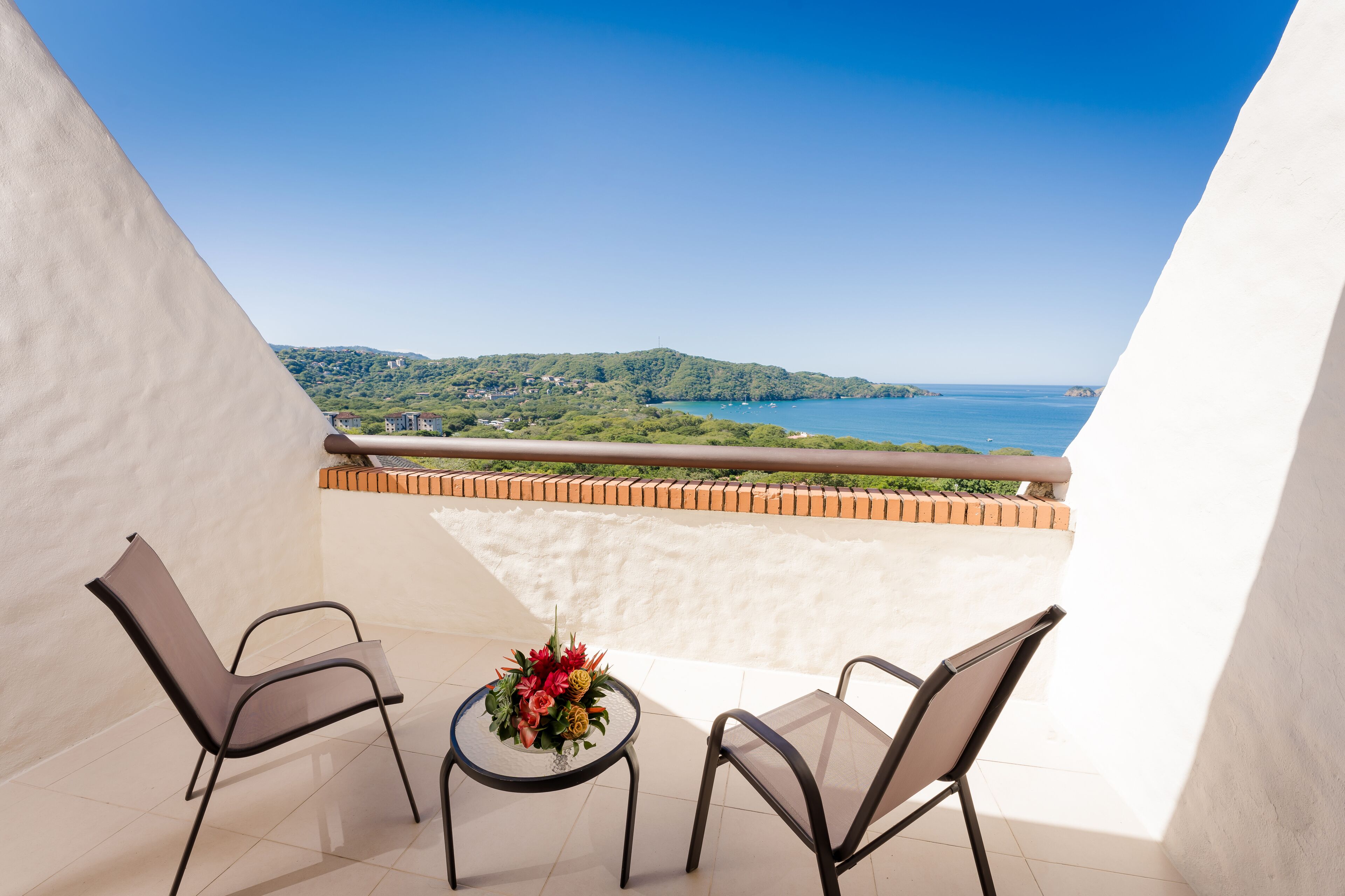 A balcony with two chairs and a small table with a flower arrangement, overlooking a coastal landscape.