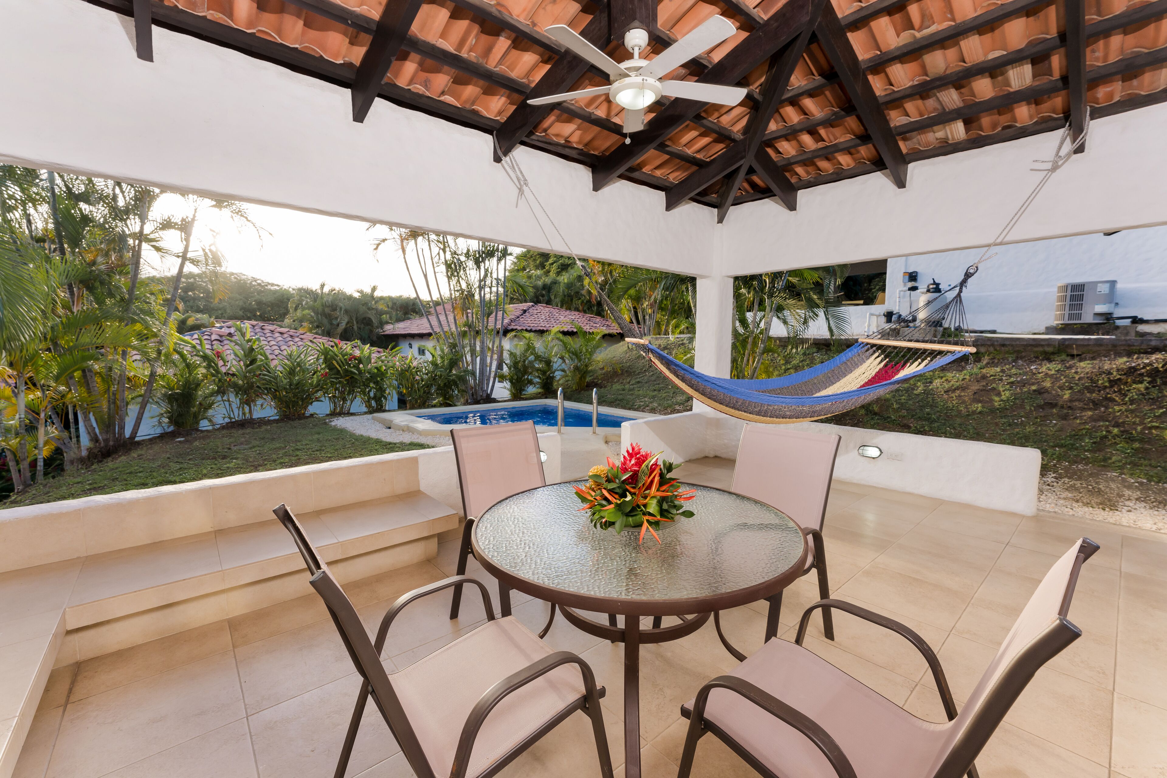 Un patio techado al aire libre con una mesa redonda y sillas, una hamaca y una piscina al fondo.