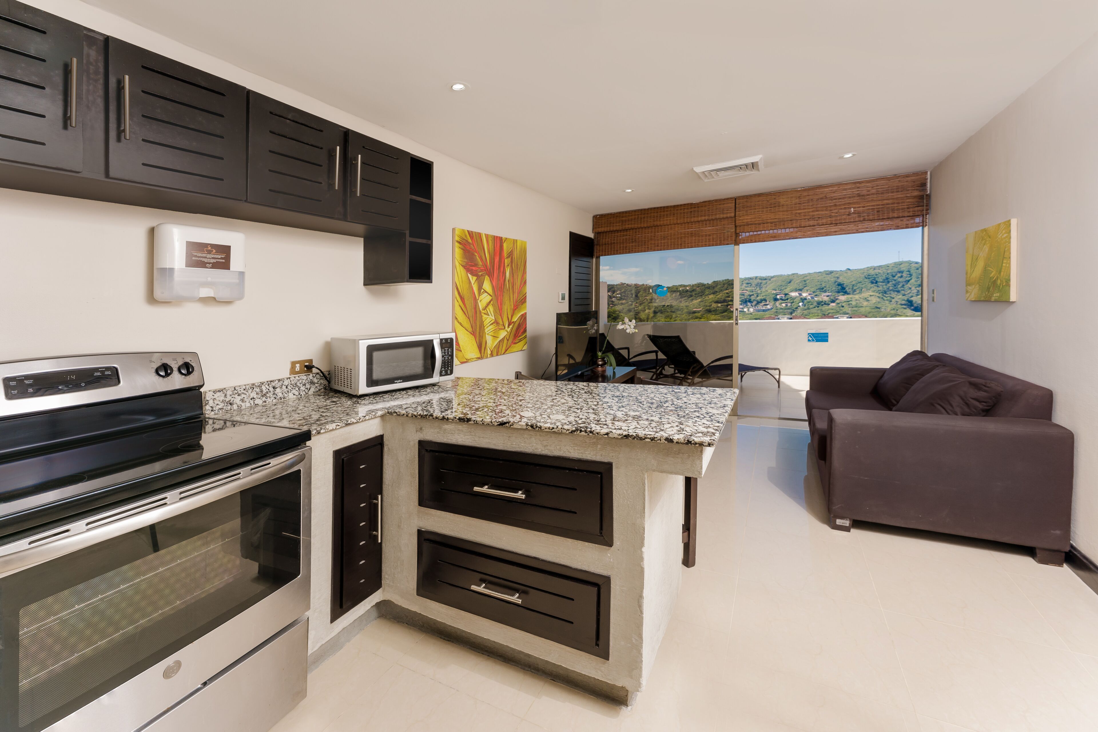 A modern kitchen with a built-in oven, microwave, and granite countertop. There is a seating area with a couch and a view of the landscape.