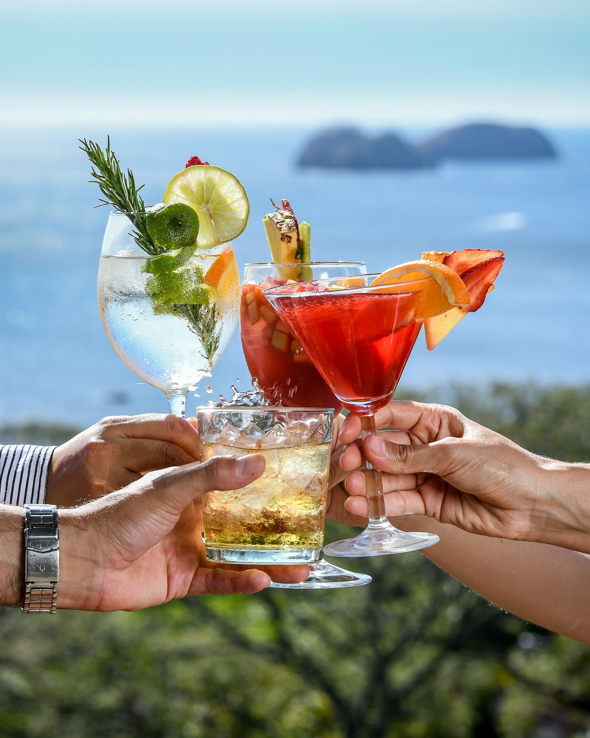 Trois cocktails, chacun agrémenté d’une garniture, tenus dans des mains, avec un paysage pittoresque en arrière-plan.