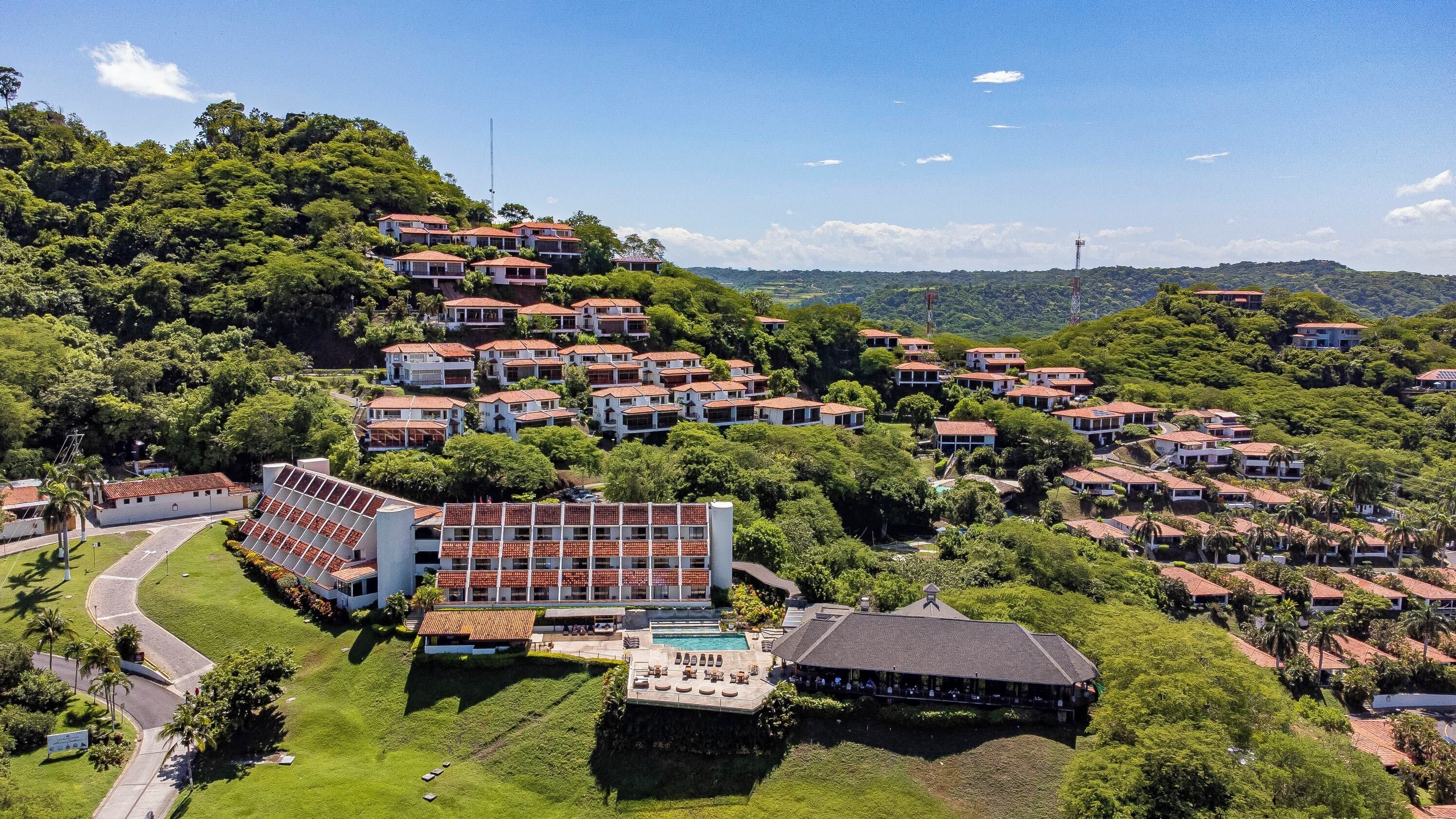 An aerial view of a residential area with multiple houses, a swimming pool, and a large building with a sloped roof.