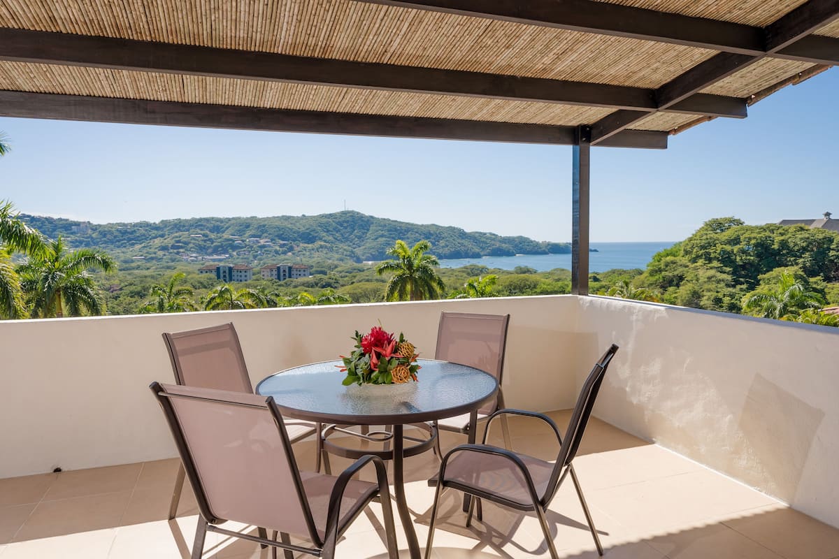 A round table with chairs on a balcony overlooking a scenic view of hills and a body of water.
