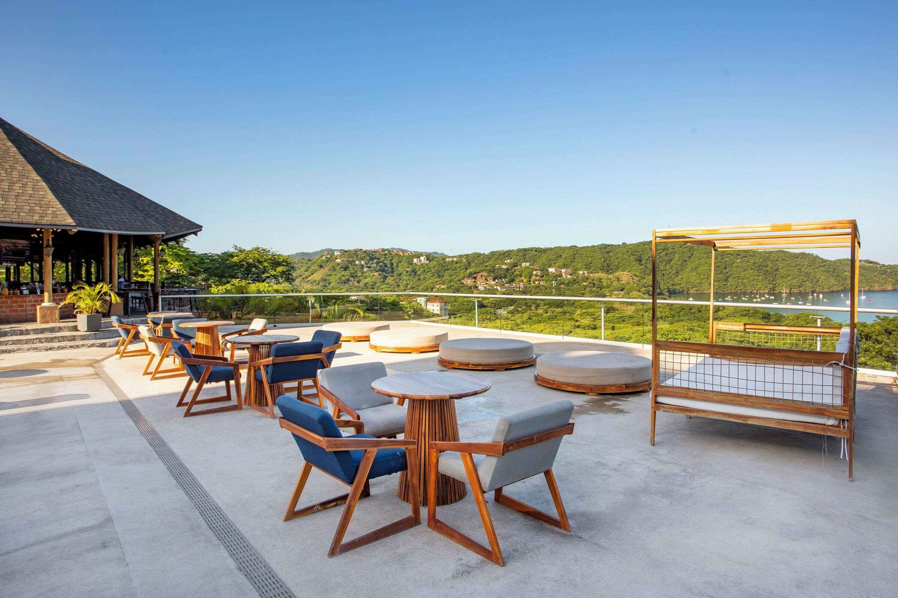 Een terras met tafels en stoelen, een houten pergola en uitzicht op een meer en heuvels.