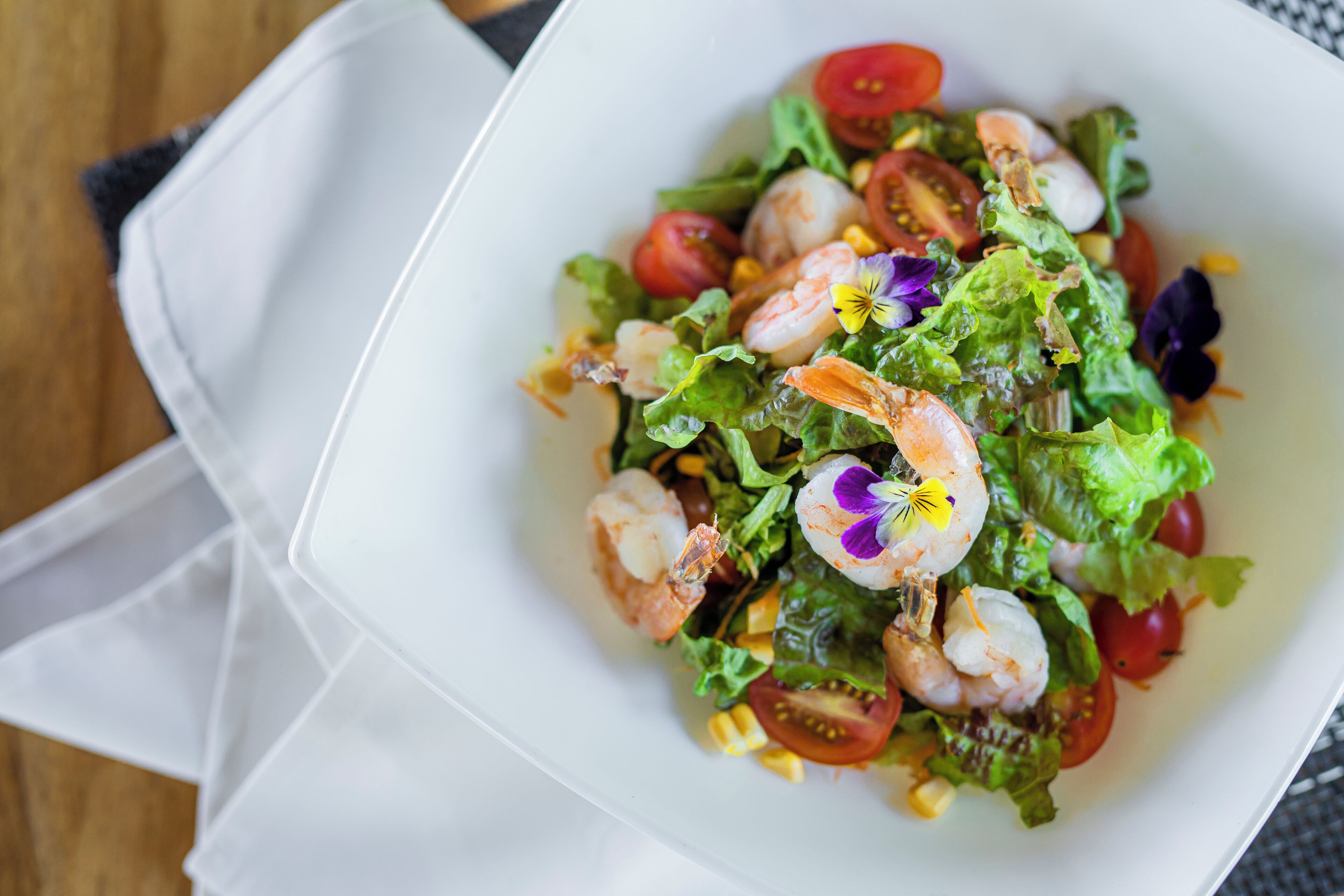 Un tazón blanco con ensalada de camarones, lechuga, tomates, maíz y flores decorativas.