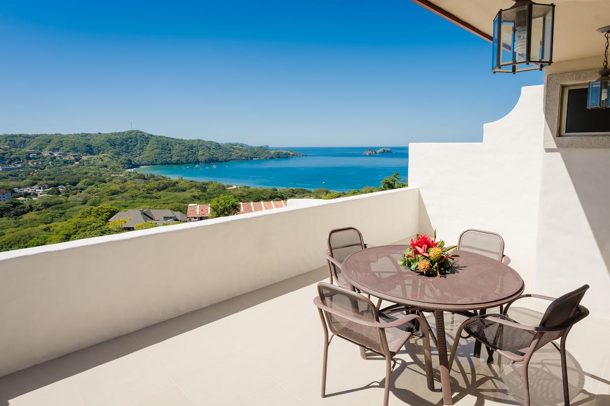 A balcony with a round table and chairs, overlooking a coastal landscape with hills and the sea.
