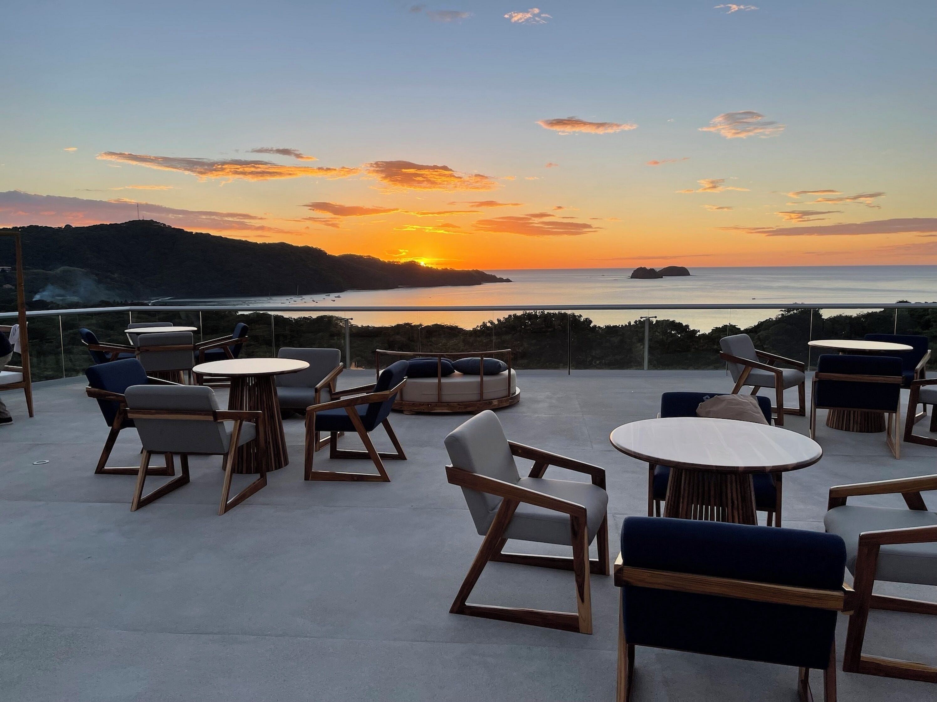 An outdoor seating area with round tables and chairs, overlooking a body of water and a sunset.