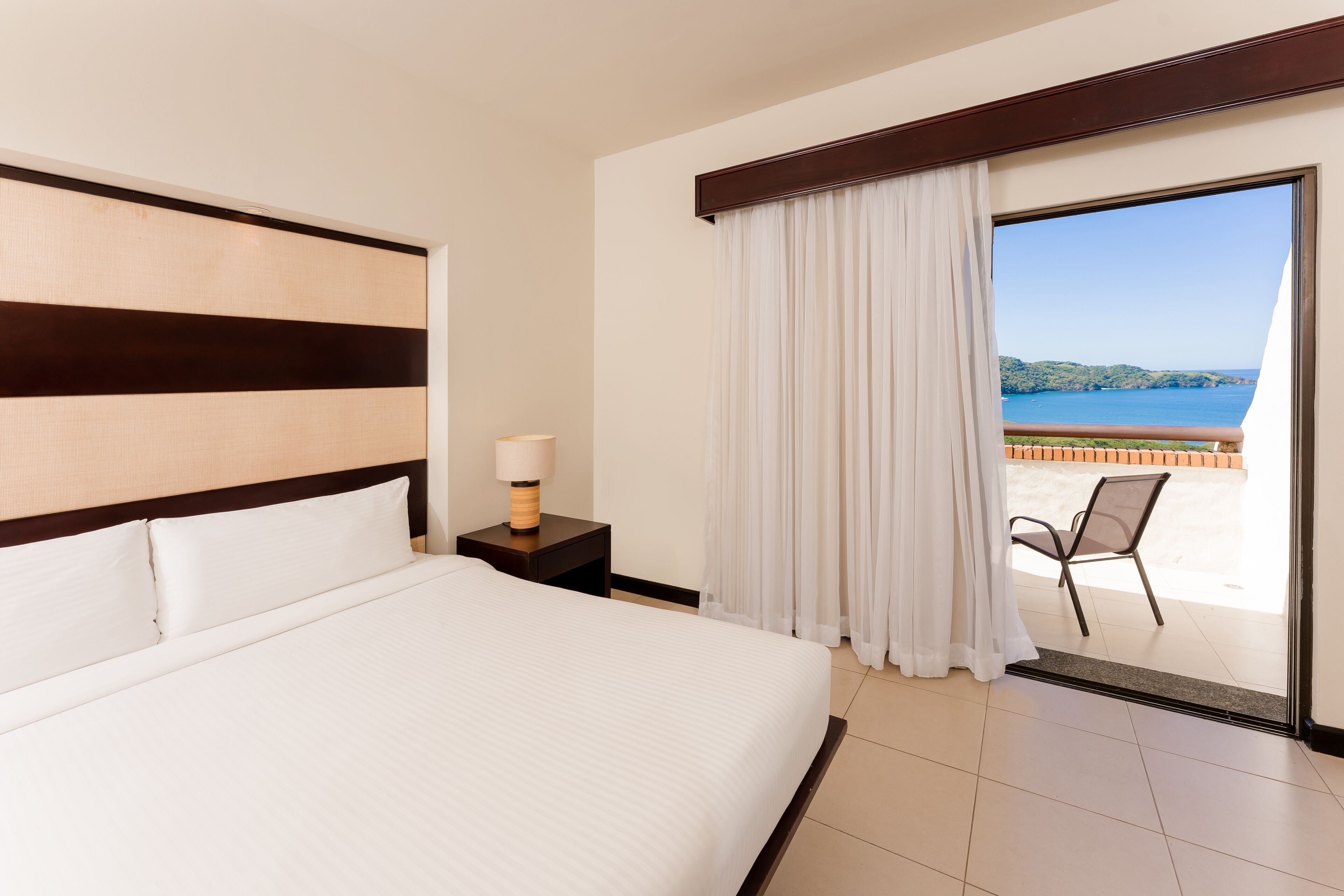 A hotel room with a large bed, a nightstand with a lamp, a view of the sea through a sliding glass door, and a chair by the door.