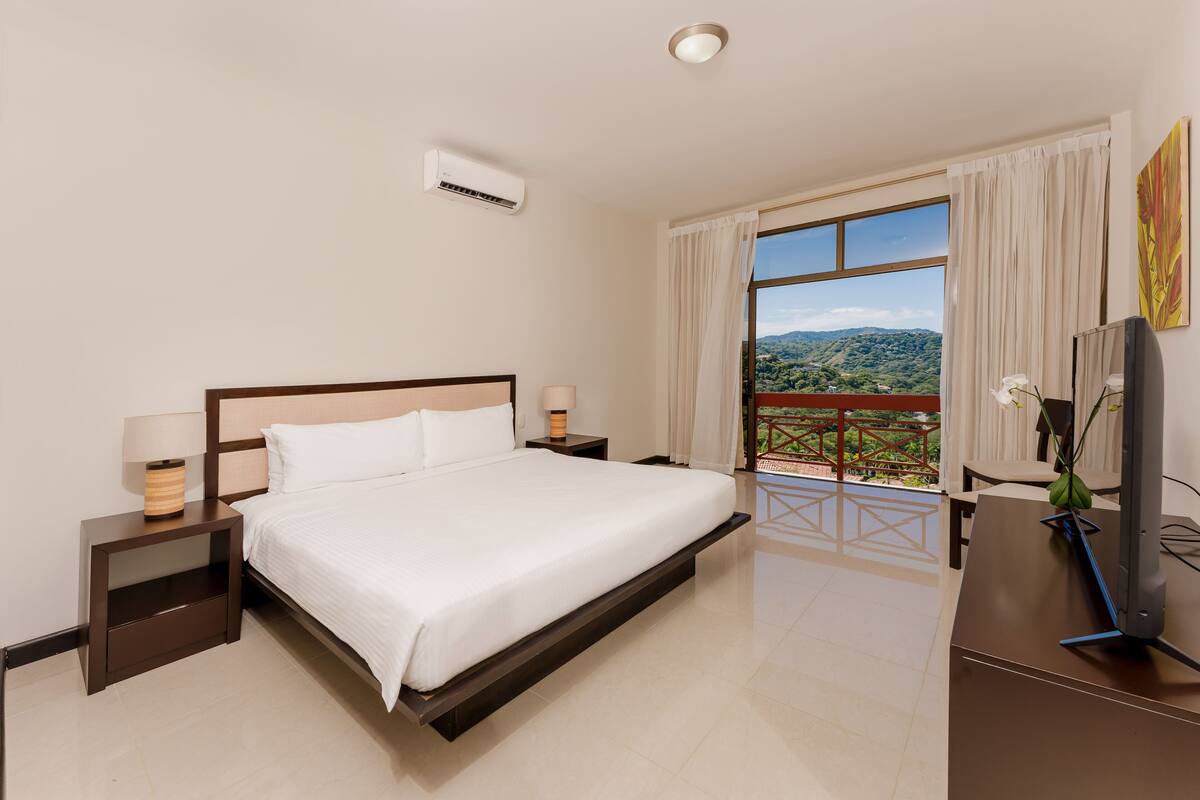 A hotel room with a large bed, a desk with a computer, a TV, and a balcony with a view of the countryside.