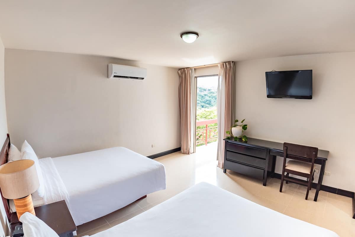 A hotel room with two beds, a desk, a chair, a TV, and an air conditioning unit.