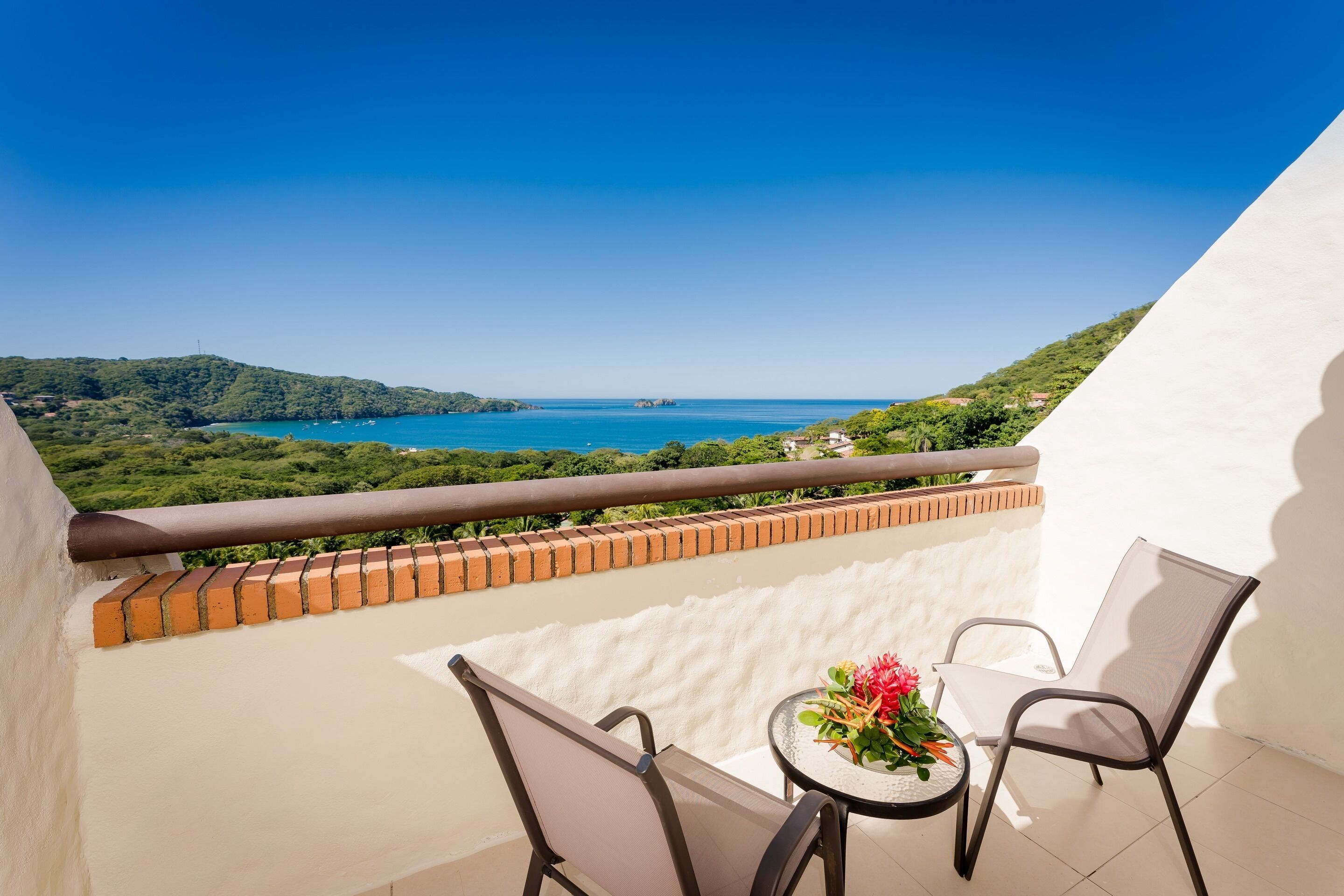 A balcony with a table and chairs, overlooking a coastal landscape with greenery and the sea.