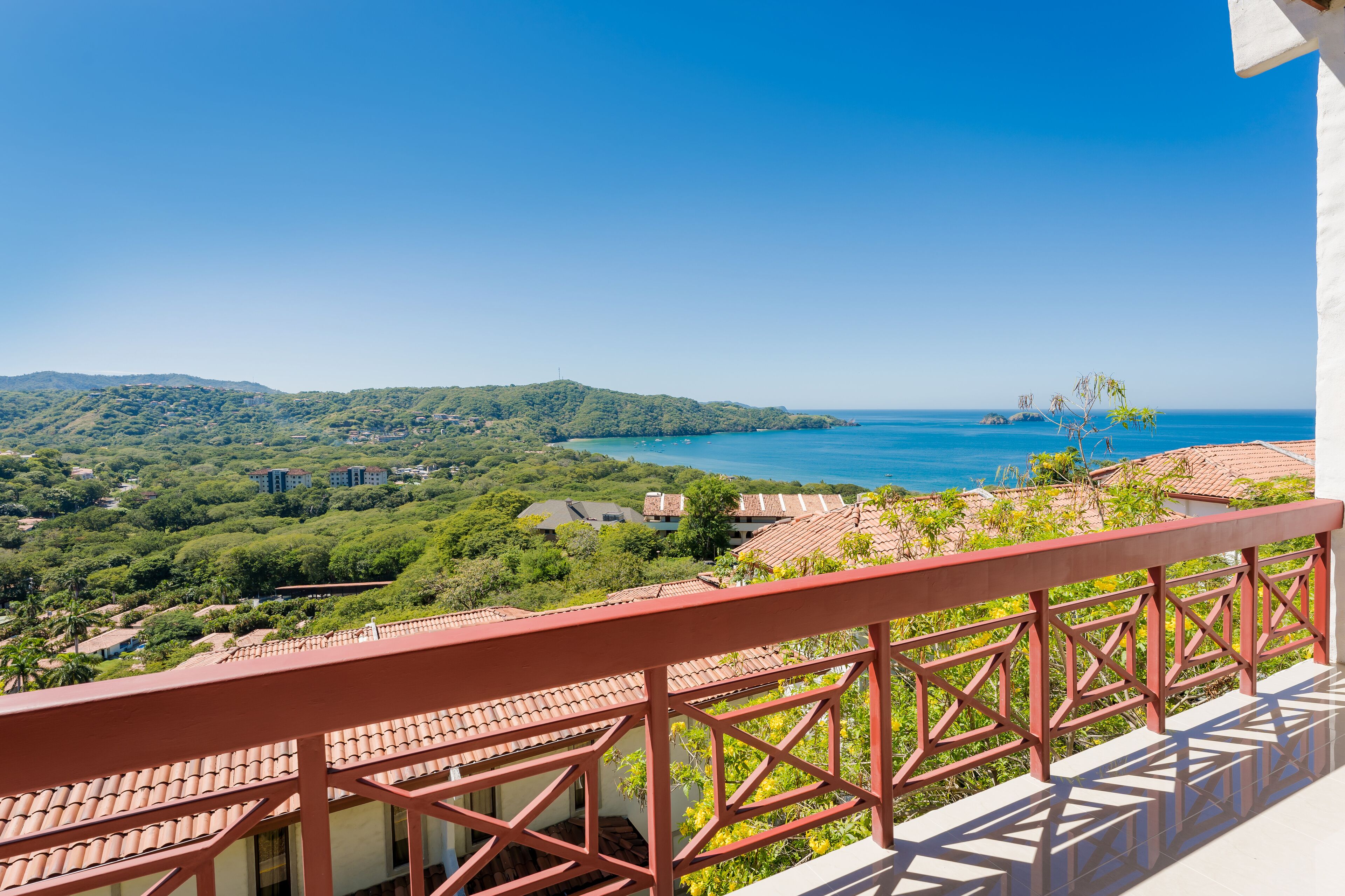 A balcony with a red railing overlooking a coastal landscape with buildings and greenery.