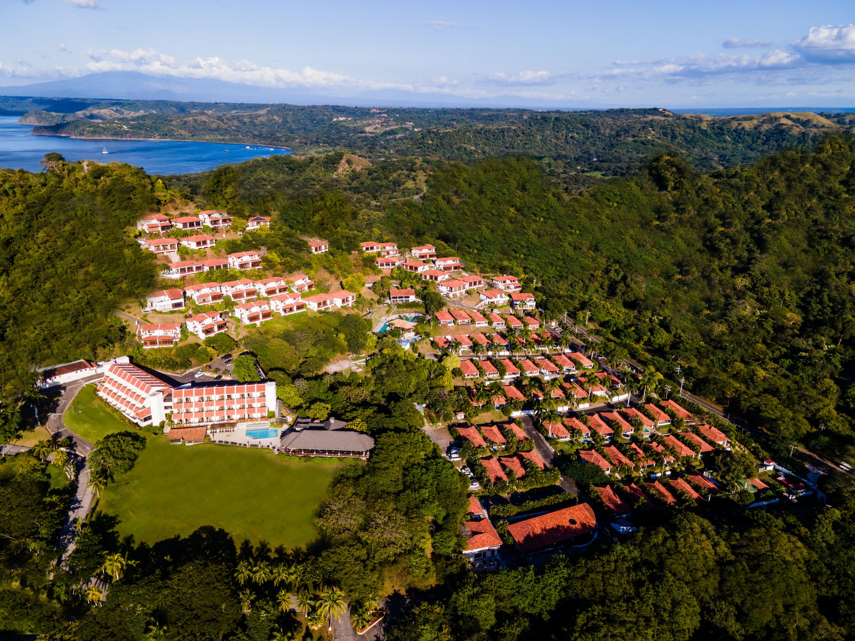 Vista aérea de un complejo turístico con varias edificaciones, una piscina y una amplia zona verde.